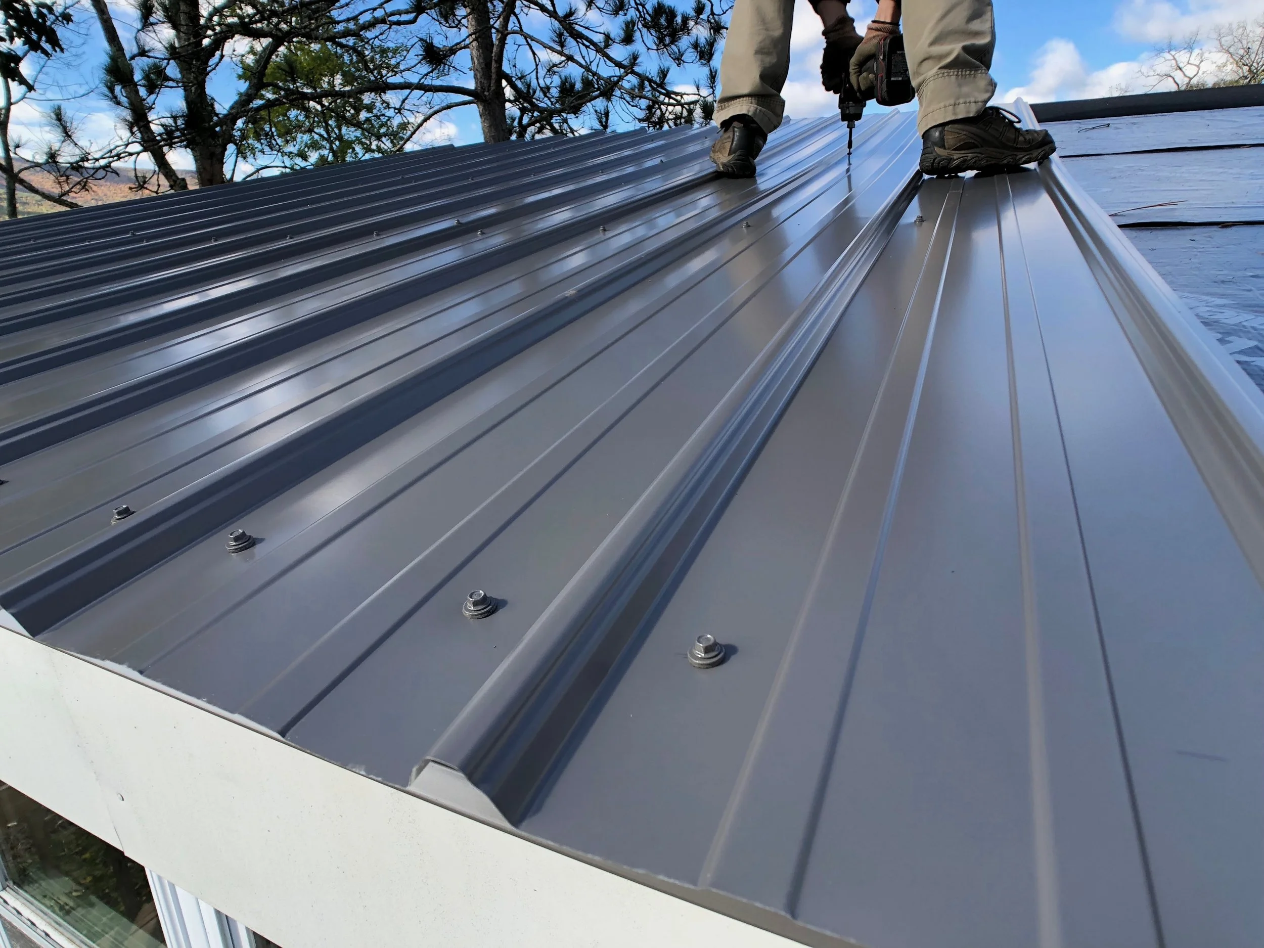 Worker installing or inspecting a shiny metal roof on a building, with trees and a blue sky in the background.