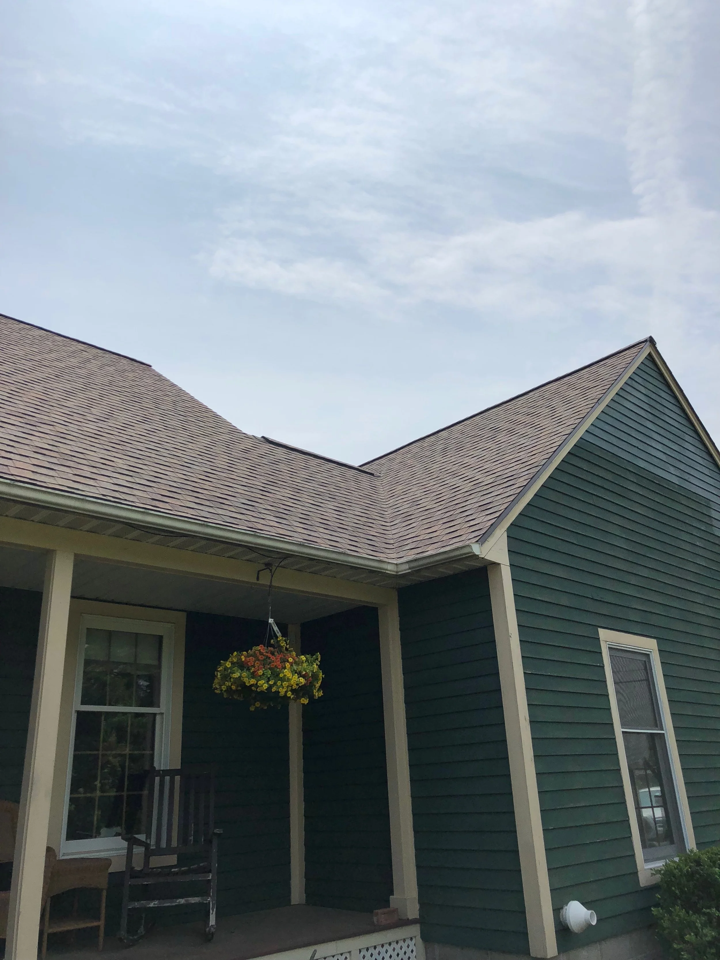 A house with dark green siding, white trim, and a brown shingle roof. A hanging flower basket is on the porch, with a black chair and a wooden chair nearby.