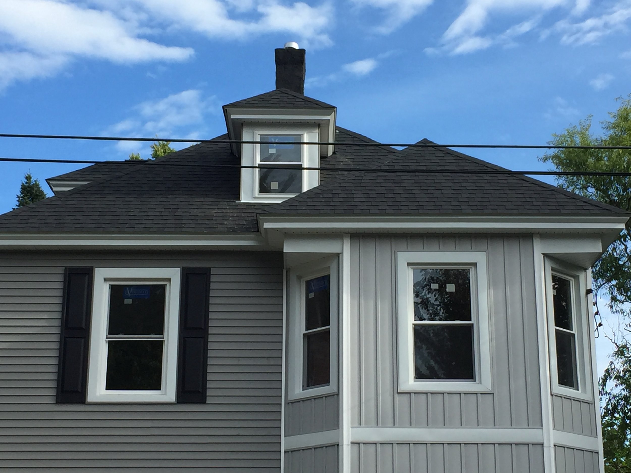Close-up of the upper part of a house with a pitched roof, dormer window, and bay window, with power lines overhead and a partly cloudy blue sky.