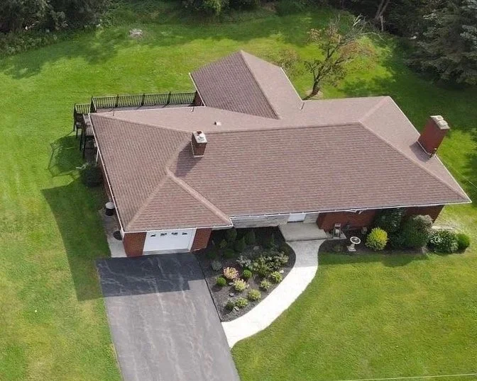 Aerial view of a single-family house with a brown tiled roof, driveway, well-maintained lawn, and surrounding trees.