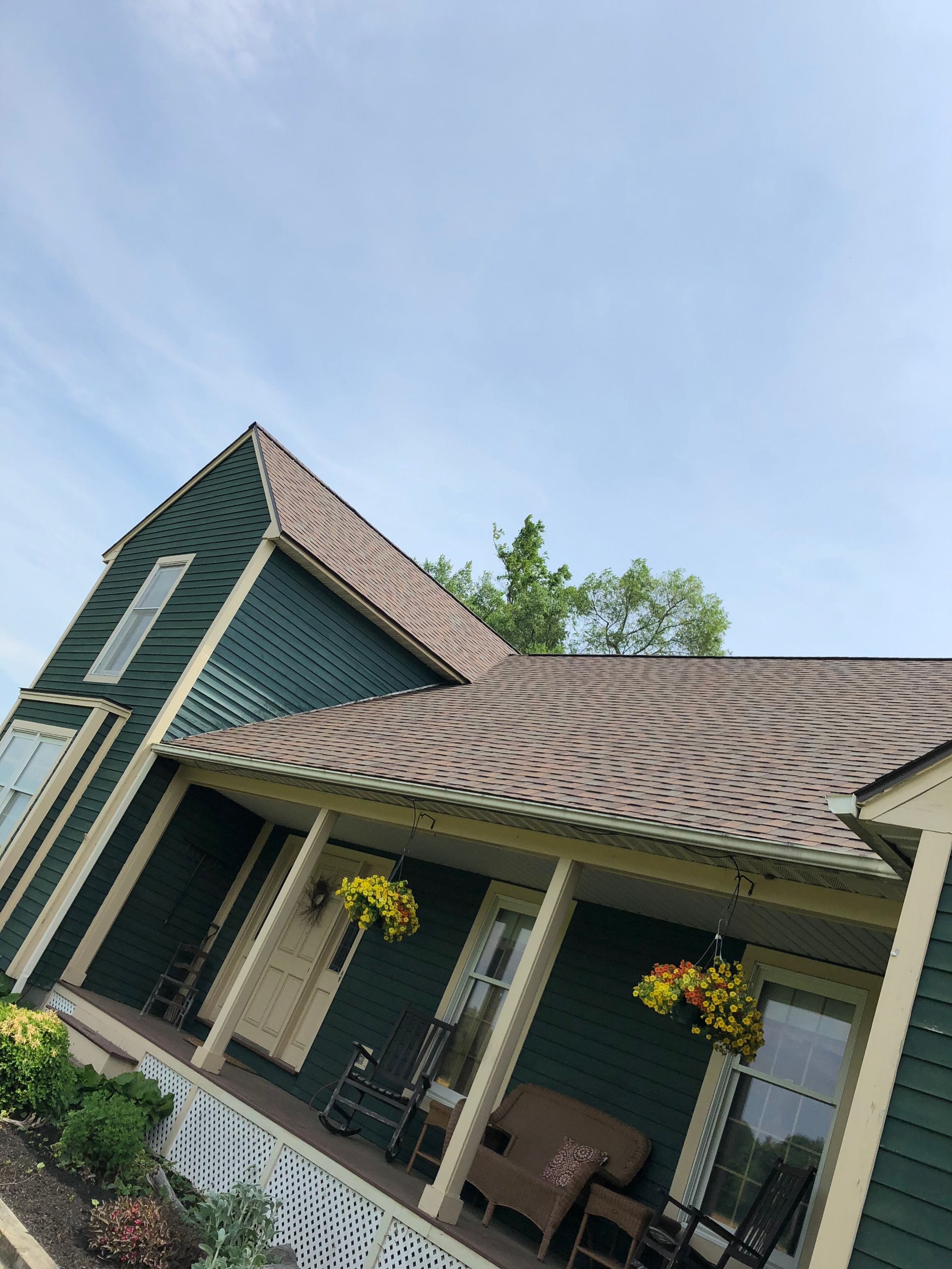 Front porch of a green house with hanging flower baskets, rocking chairs, and a porch swing, under a partly cloudy sky.