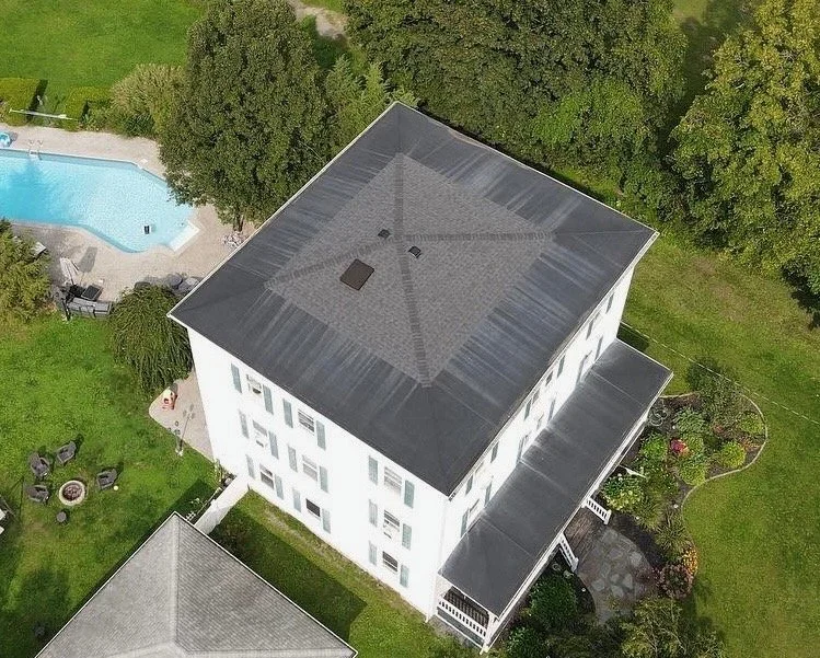Aerial view of a large white house with a dark gray roof, surrounded by green trees and lawn, with a swimming pool in the backyard.