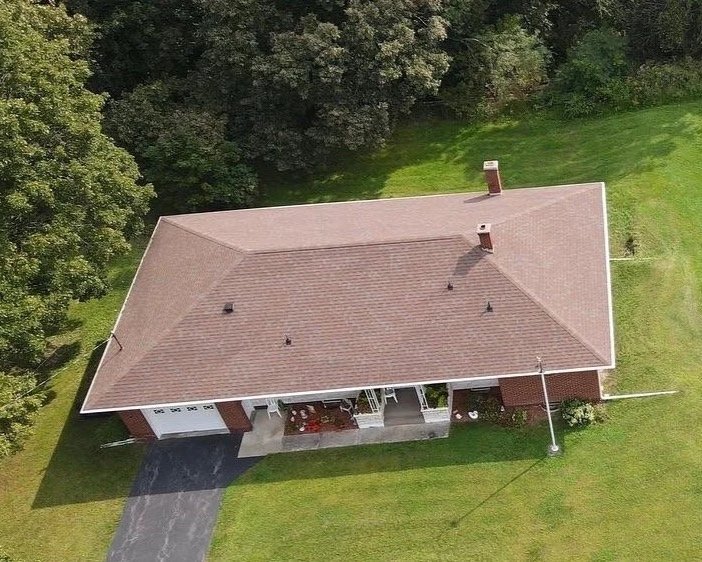 Aerial view of a single-story house with a brown roof, a driveway leading to a garage, a porch with outdoor furniture, and surrounded by green lawn and trees.