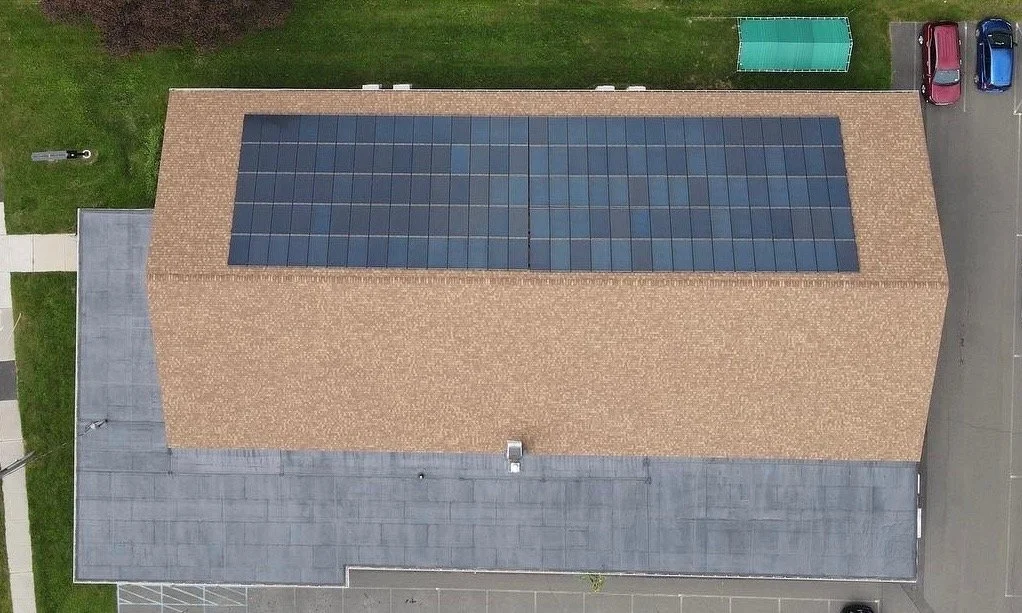 An aerial view of a building with a roof covered in solar panels, surrounded by parking spaces and some green areas.