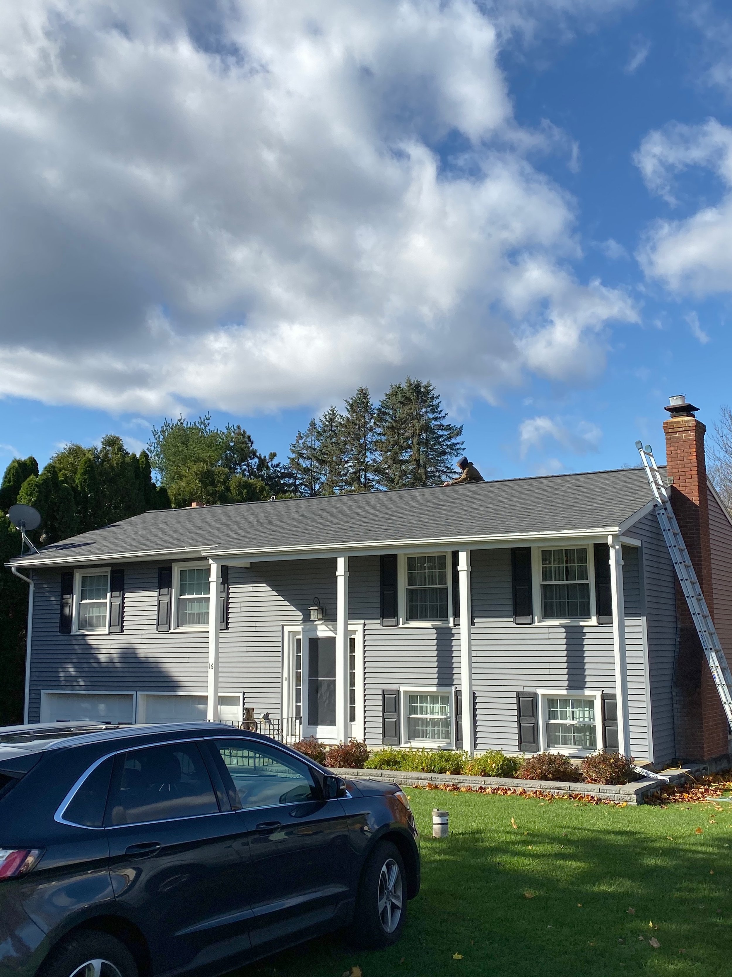 A two-story house with grey siding, black shutters, and a brick chimney. A person is working on the roof near the chimney, with a ladder leaning against the house. There is a car parked on the grass in front of the house and a landscaped garden bed w
