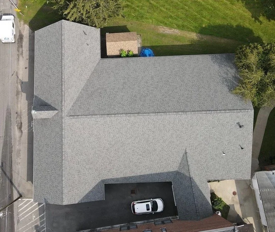 Top-down aerial view of a house with a gray roof, driveway with a white car, small shed, and surrounding trees and grass.