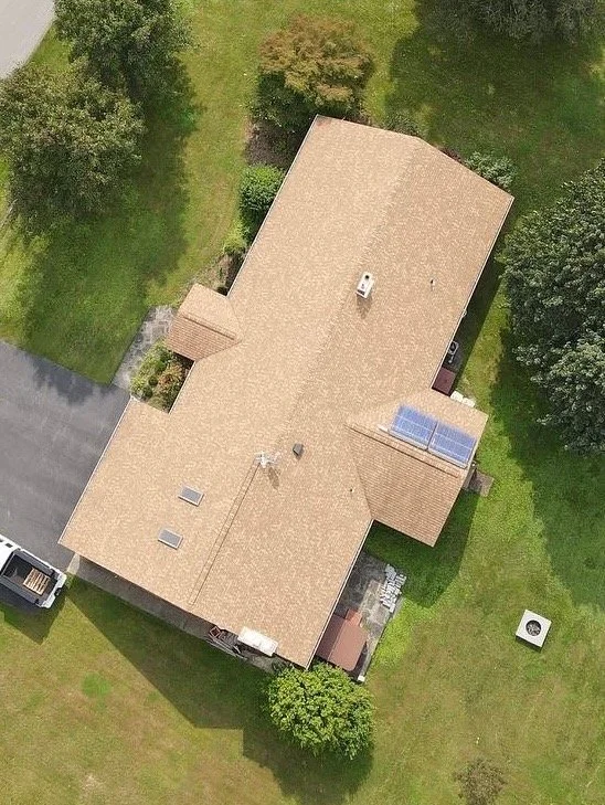 Aerial view of a house with a tan roof, surrounded by green grass and trees, and a driveway with a vehicle parked on it.