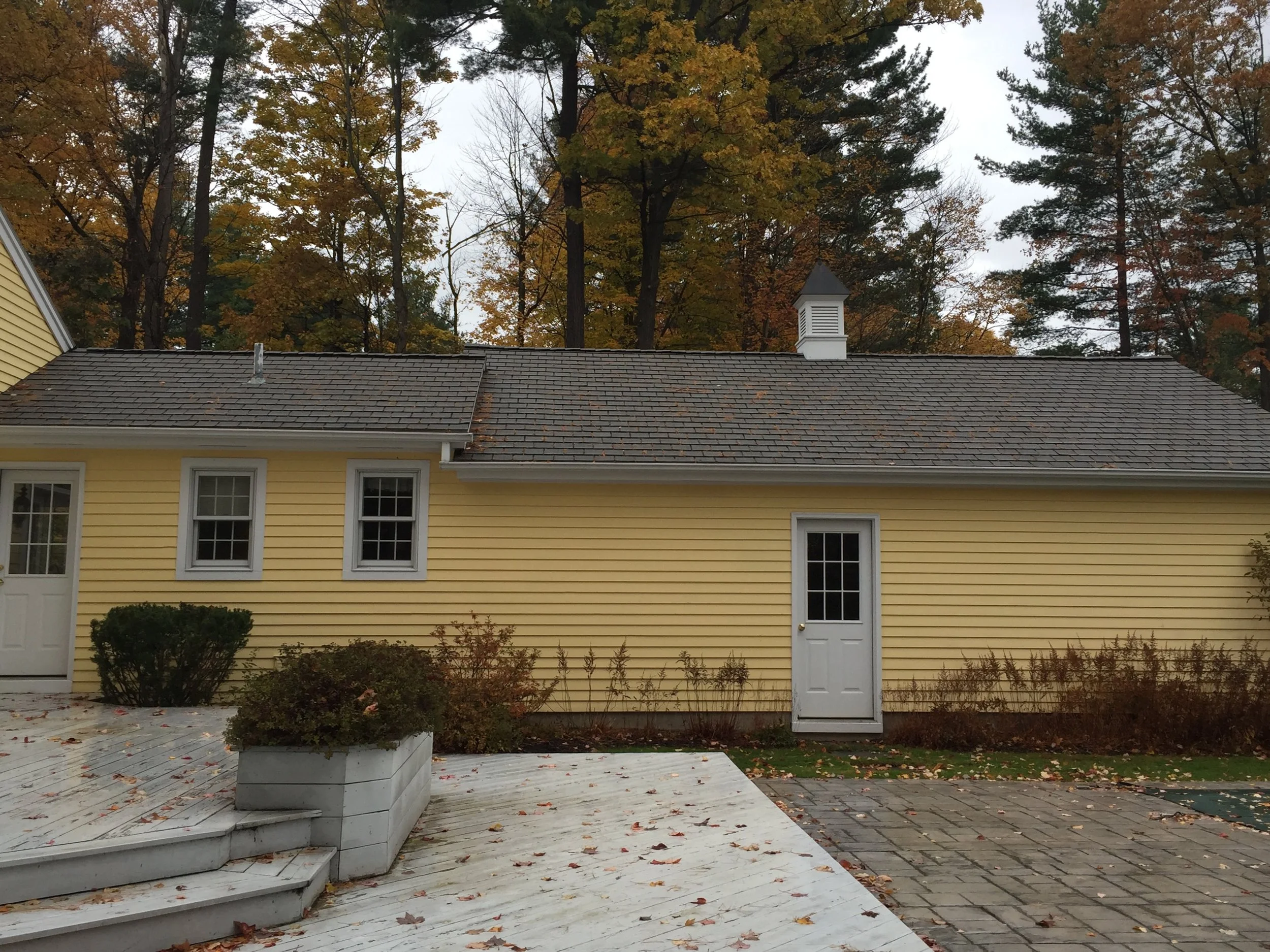 A yellow house with a gray shingle roof, three small windows, and a white door. There are some bushes and fallen leaves in the yard, with a pathway and steps leading to the house. Tall trees with autumn leaves are in the background.