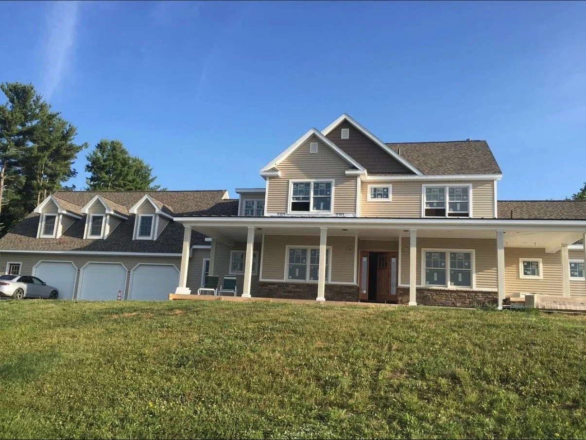 A large two-story house with beige siding and white trim, featuring a front porch with white columns, multiple windows, a gabled roof, and an attached three-car garage. A white car is parked near the garage, and there is a grassy lawn in front of the