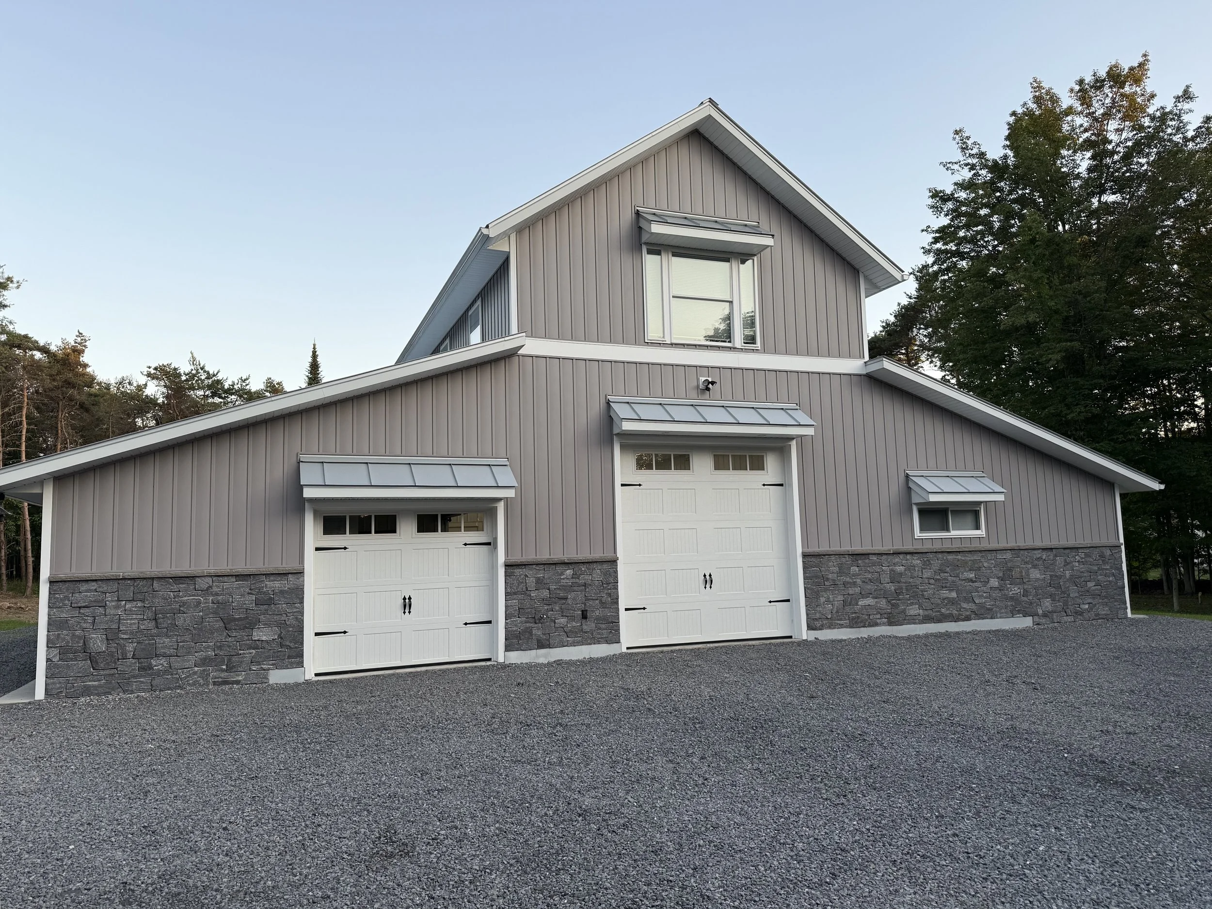 A modern two-story house with a gray exterior, white trim, and stone accents, featuring two garage doors and surrounded by trees.