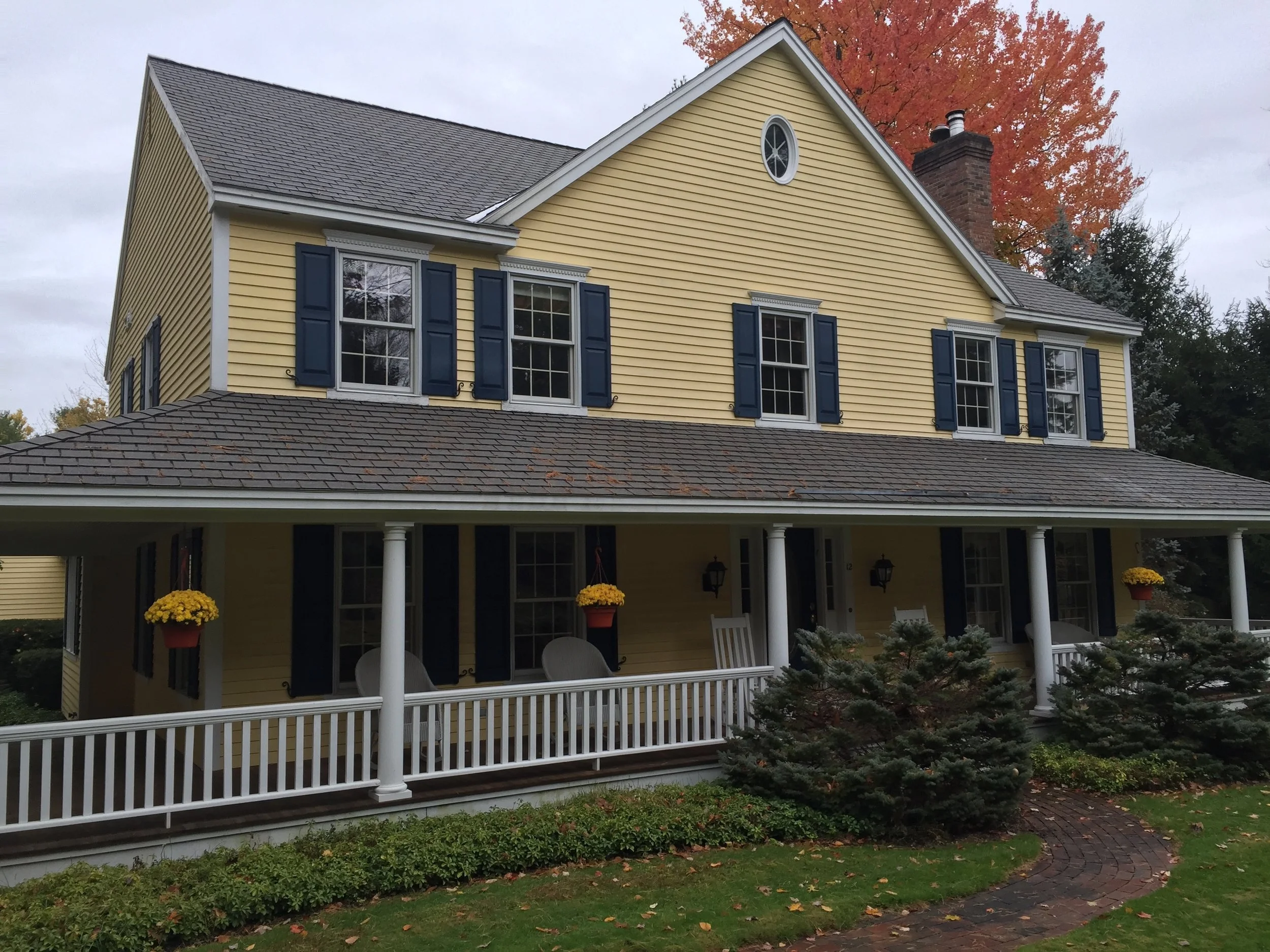 Yellow two-story house with navy shutters, a gray shingled roof, and a front porch with white railings, hanging flower pots, and yard landscaping, with fall foliage in the background.