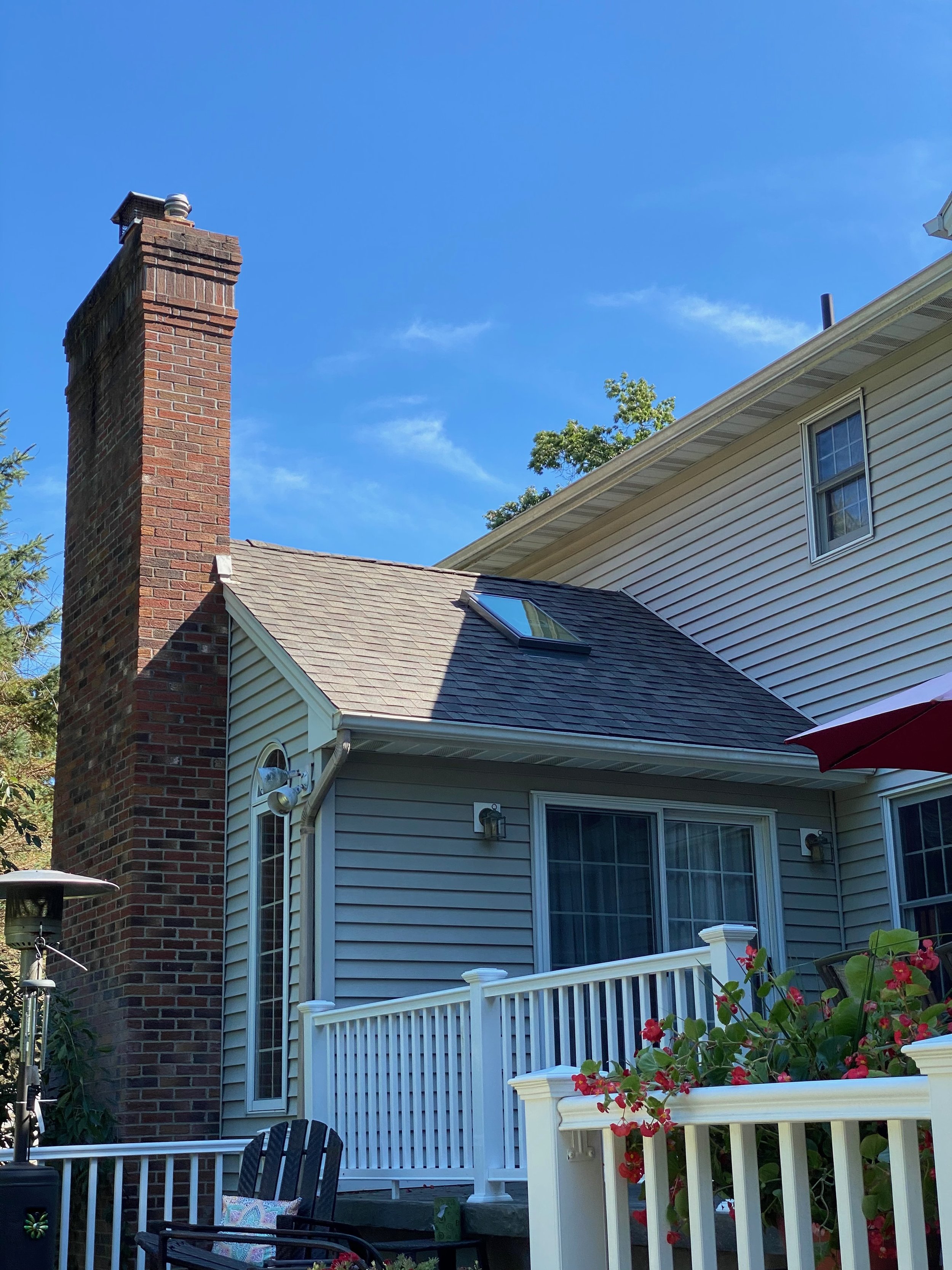 View of the backyard of a house with a brick chimney, gray sliding door, white railing, a patio chair, and a red umbrella, with partly cloudy blue sky.