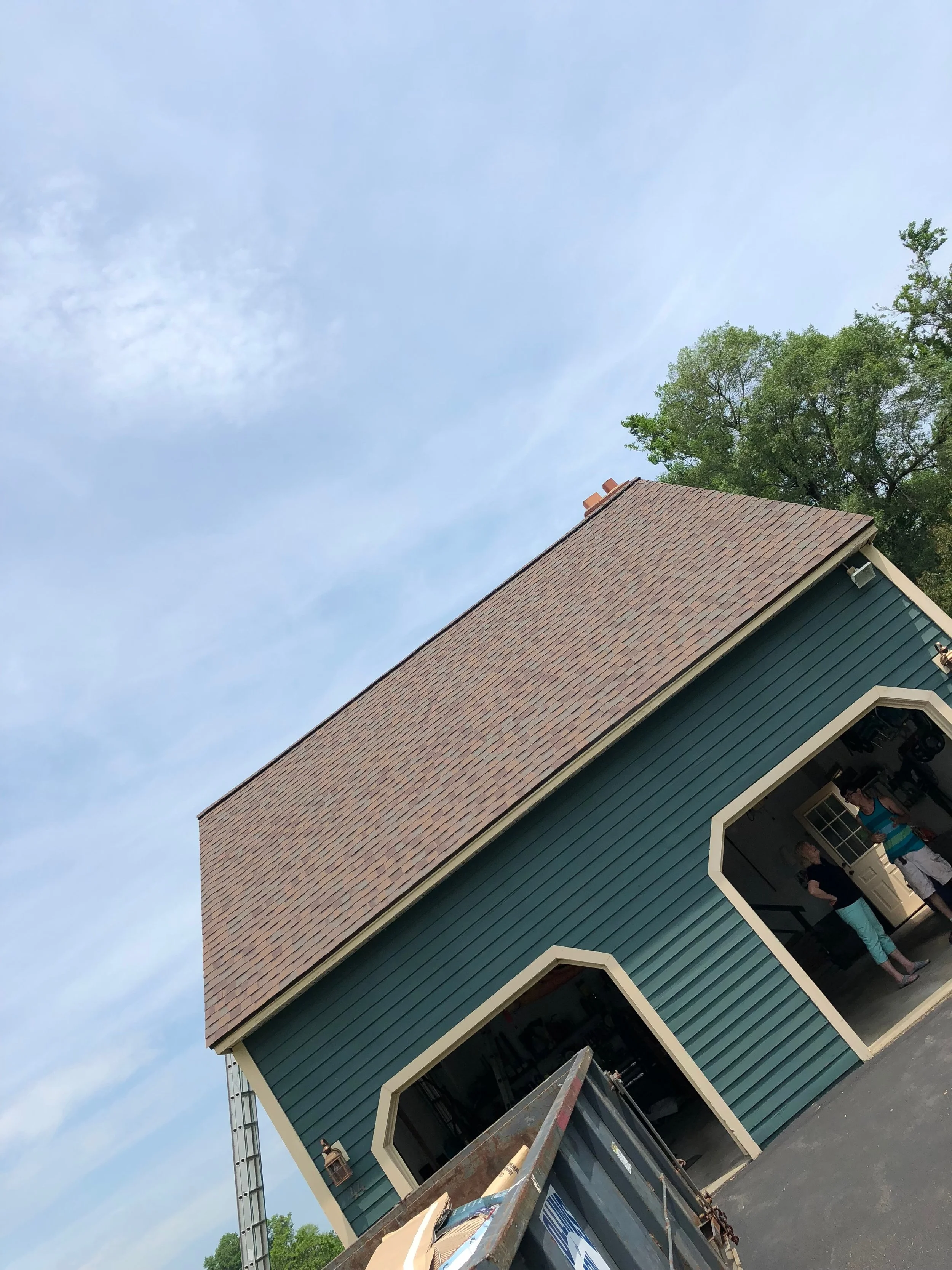 A person standing inside a garage of a teal-colored house with a brown roof, with a ladder leaning against the house and a truck parked outside.