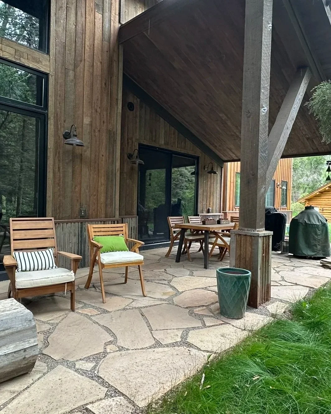 A rustic outdoor patio with wooden chairs, a table, and a potted plant on a stone floor, attached to a wood-paneled house in a forested area.
