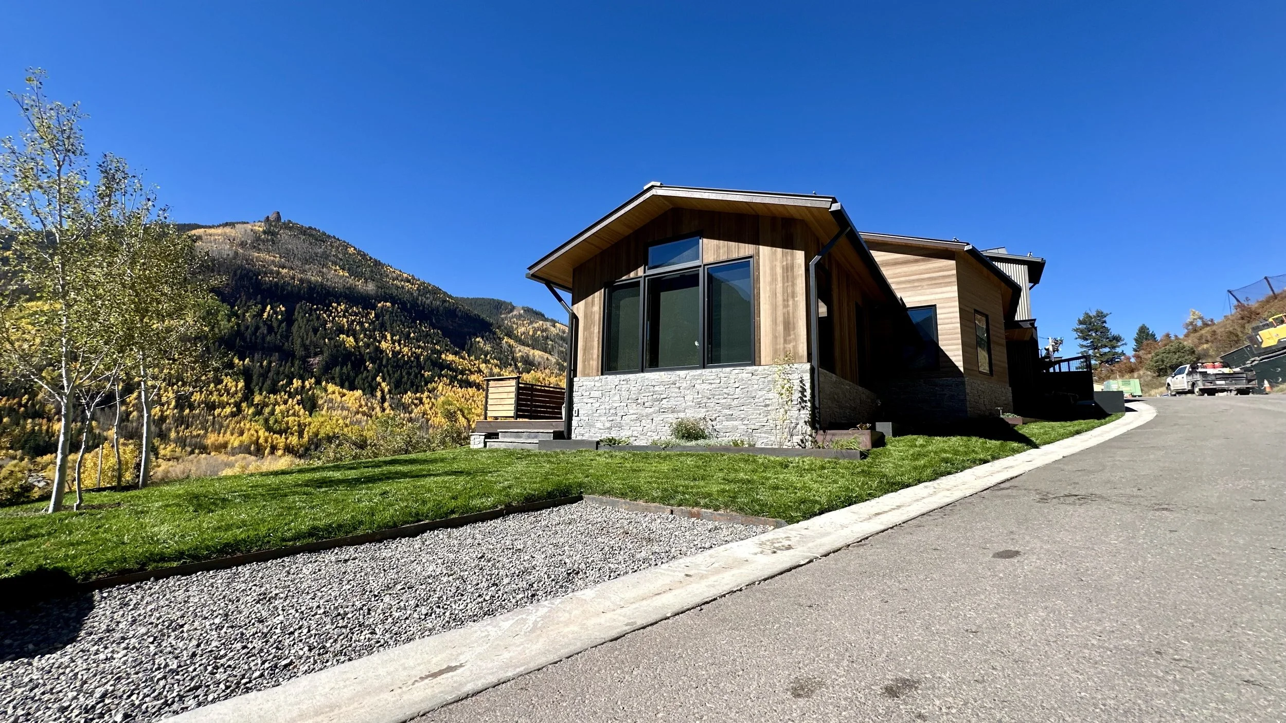 Modern house with large glass windows and wooden siding on a sloped driveway, surrounded by green lawn and trees, with a backdrop of mountains with autumn foliage and a clear blue sky.