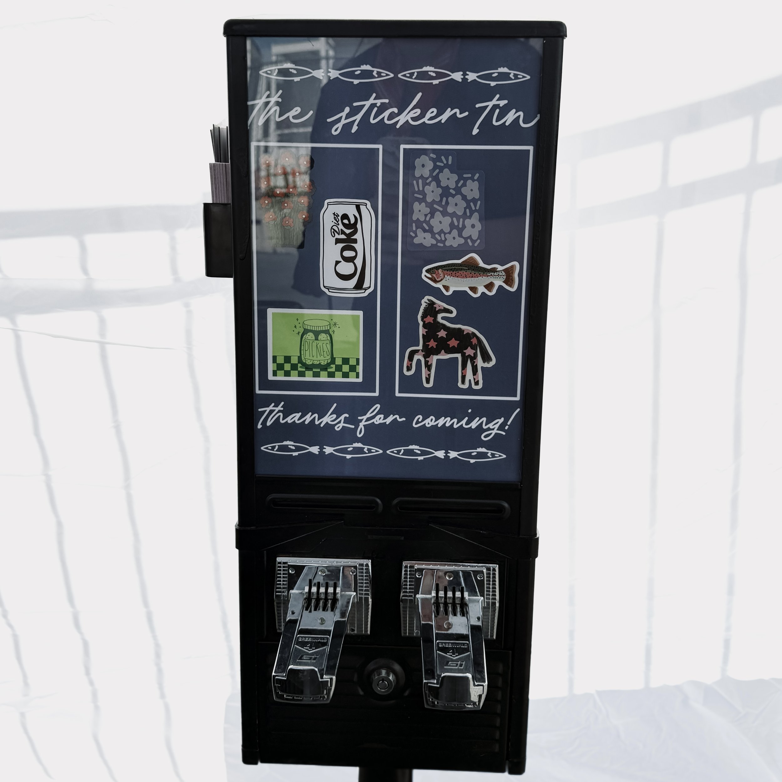 A vending machine for fish-shaped snacks with a sticker of a fish and various decorative stickers, with a white background and shadows cast on the surface.