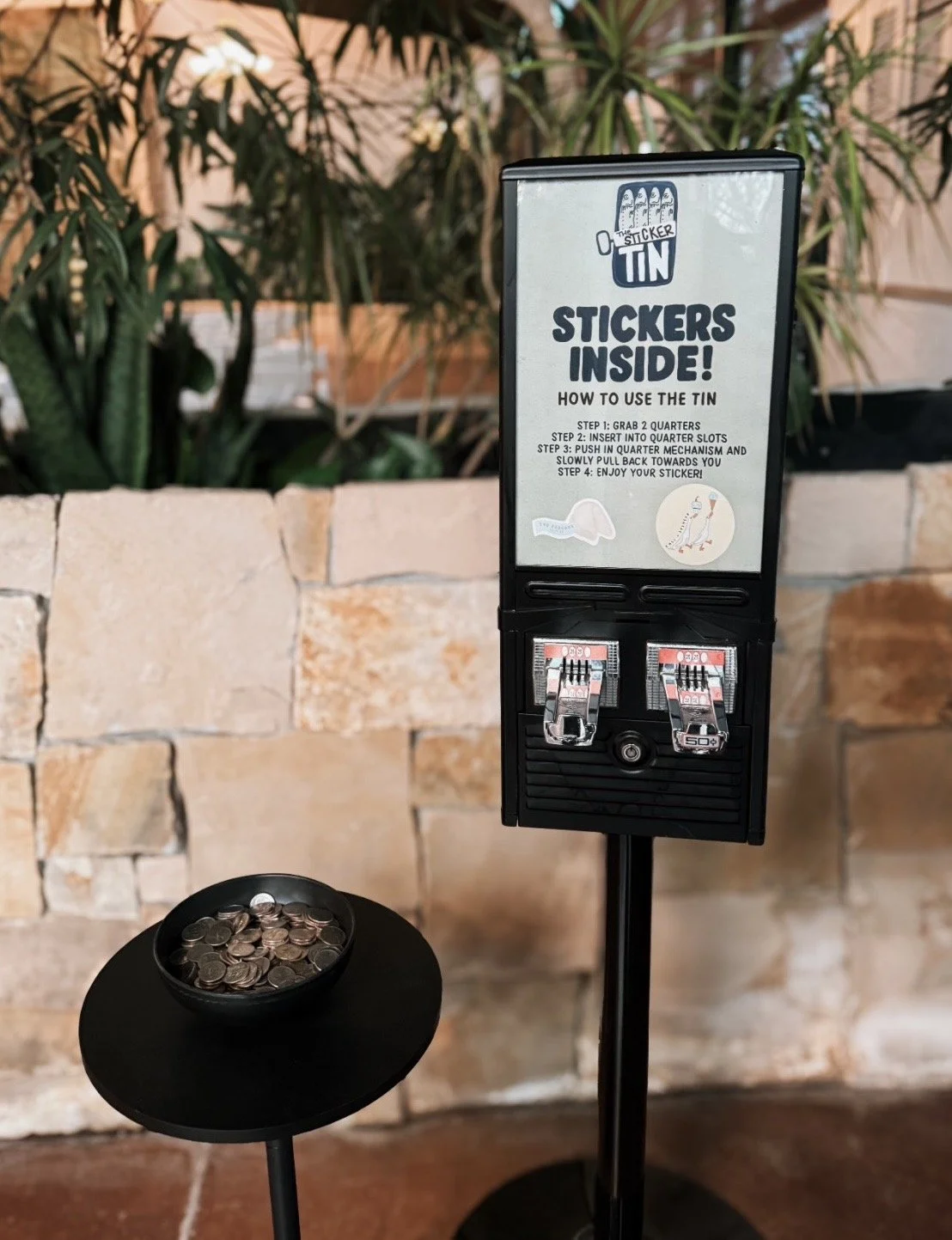 A black donation tin with instructions for using it, including steps to insert coins, push the mechanism, and collect stickers, inside a setting with a small table holding a bowl of coins in front of a brick wall background.