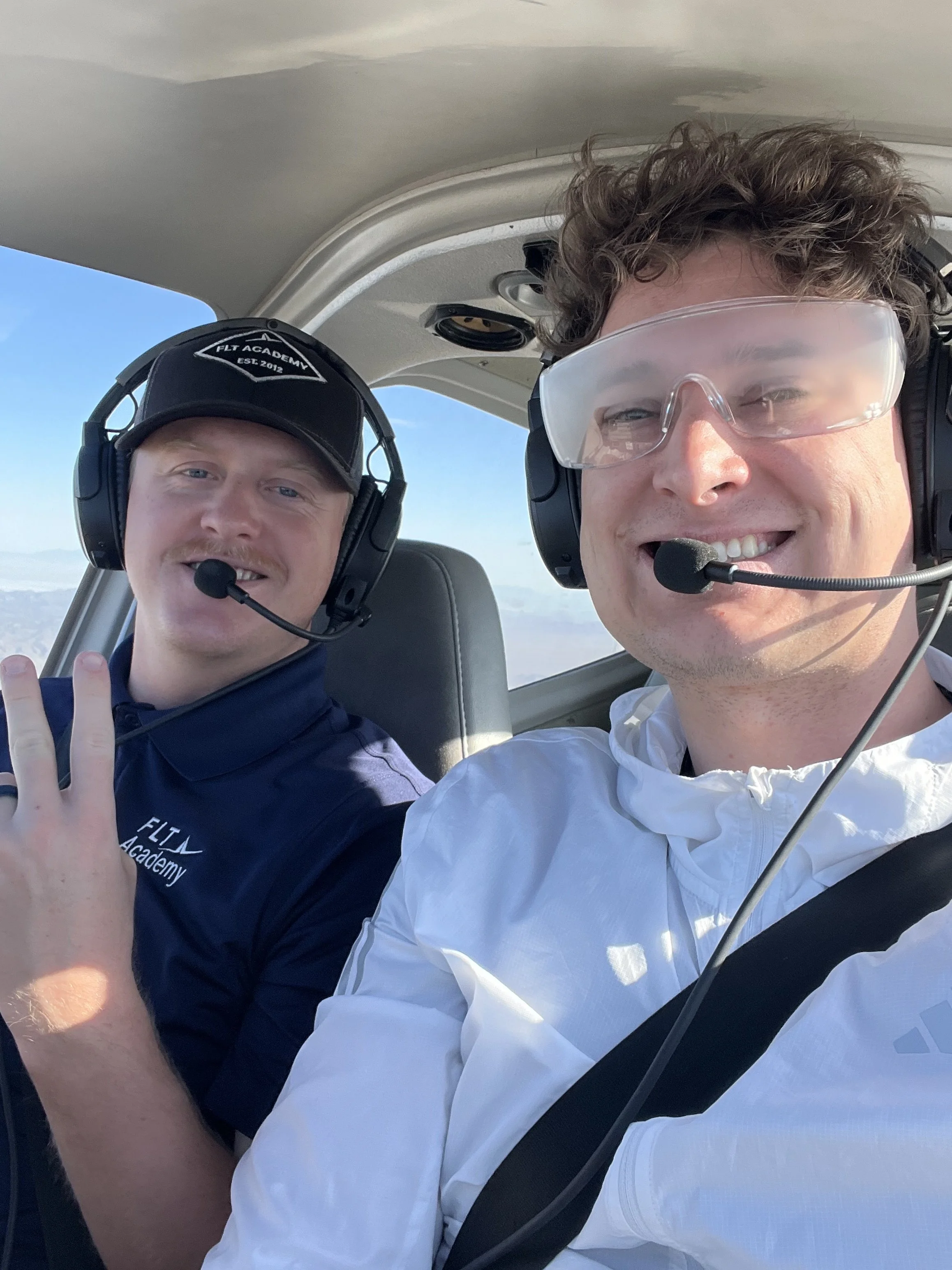 Two men inside a small aircraft, wearing headsets with microphones, smiling at the camera. One man is making a peace sign with his fingers and wearing a cap and a navy shirt. The other man is wearing clear glasses and a white jacket. Blue sky and clouds are visible outside the aircraft window.