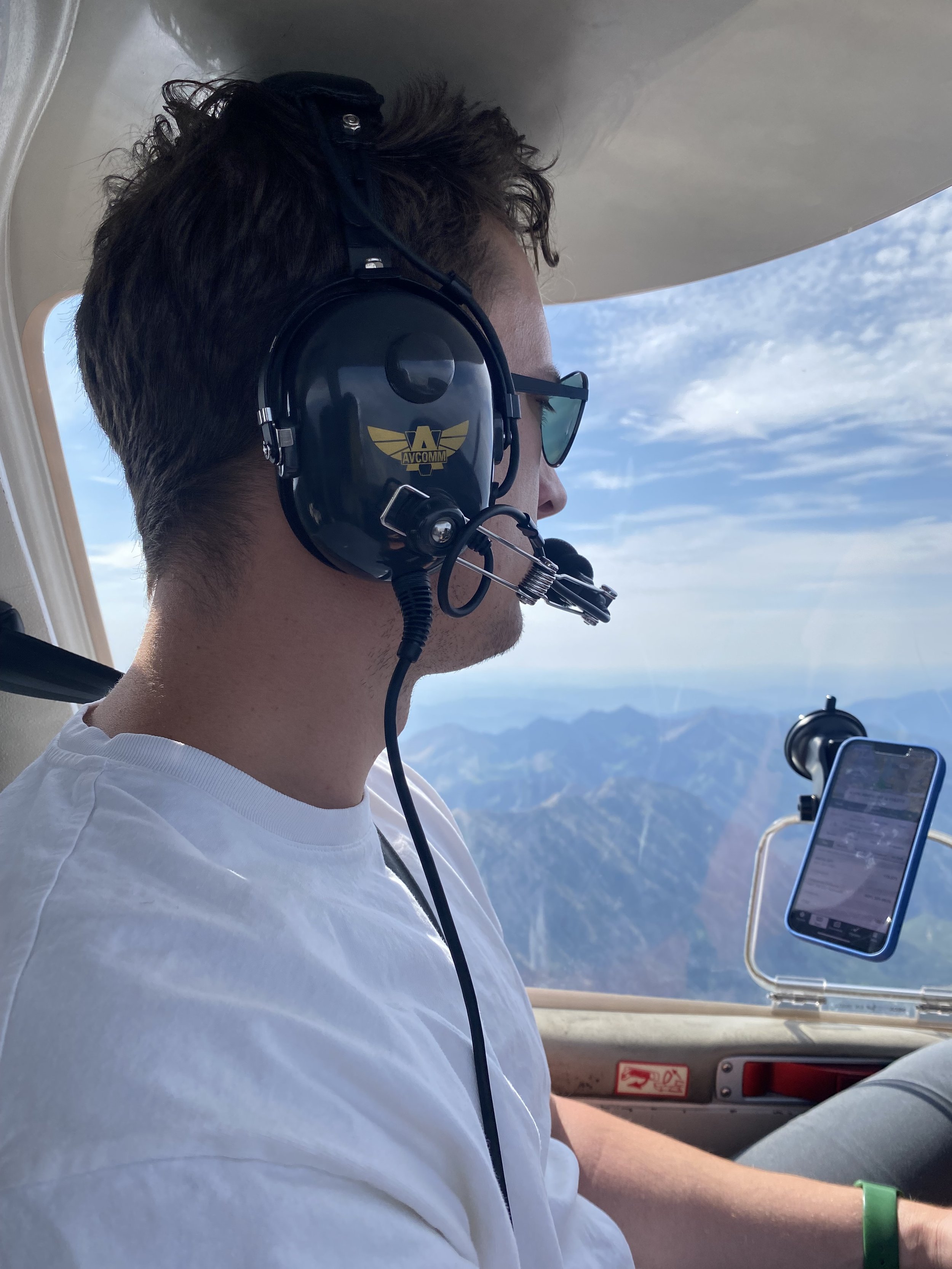 A man wearing pilot headphones and sunglasses, sitting inside an aircraft cockpit with a view of mountains and sky outside.