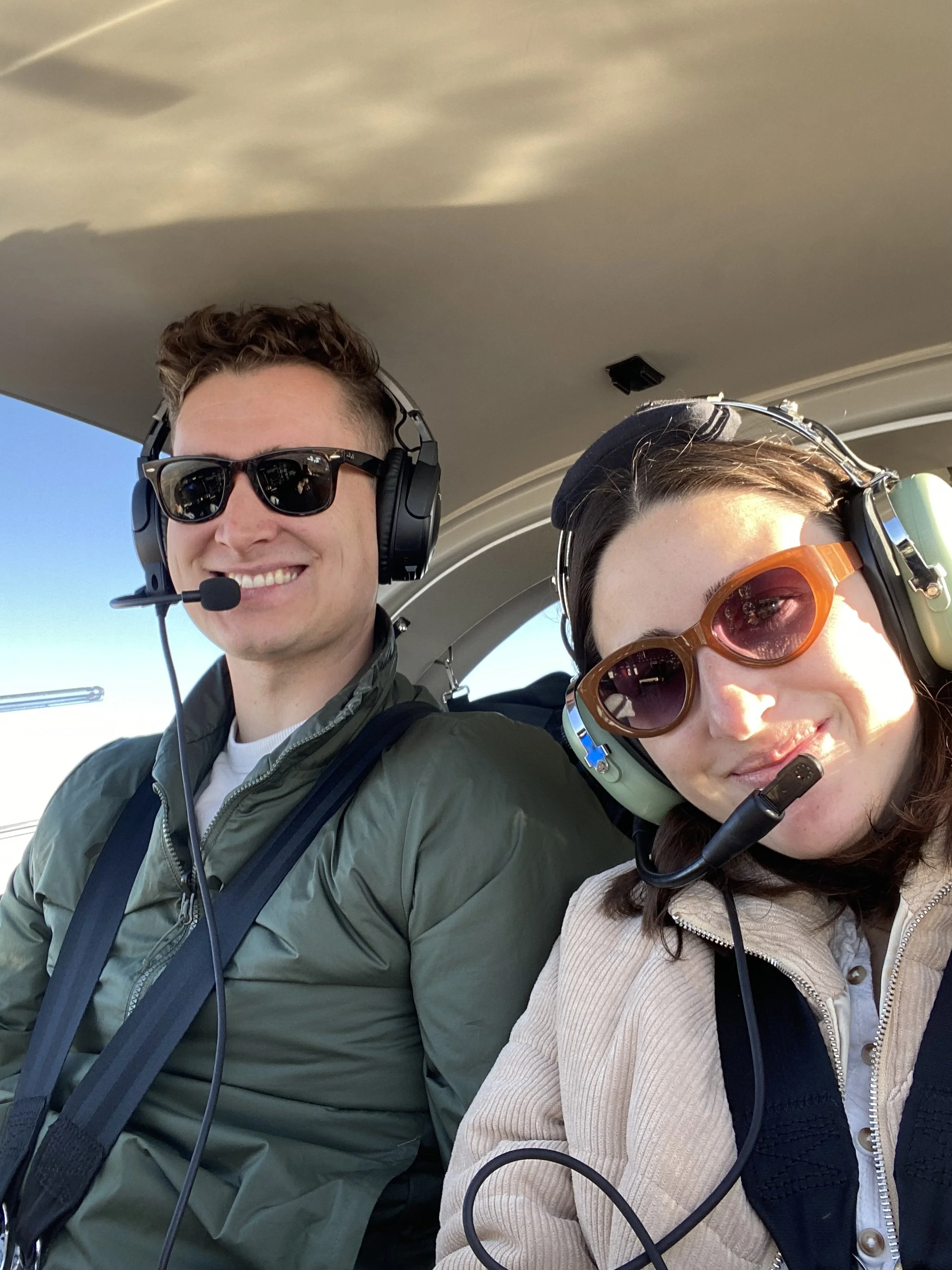 A smiling man and woman sitting in the cockpit of an airplane, wearing headsets with microphones, the man in sunglasses and green jacket, and the woman with dark brown hair and orange sunglasses.