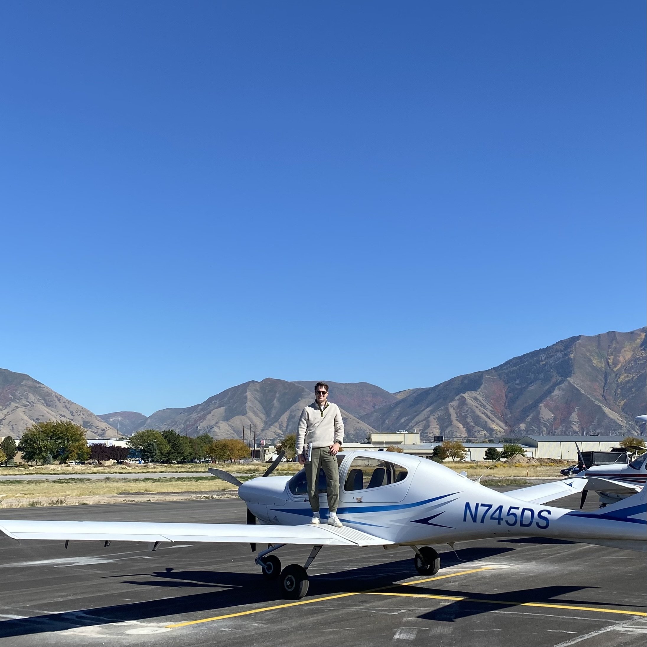 A man standing on the wing of a small white airplane, parked on a tarmac at an airport with mountains in the background and a clear blue sky overhead.
