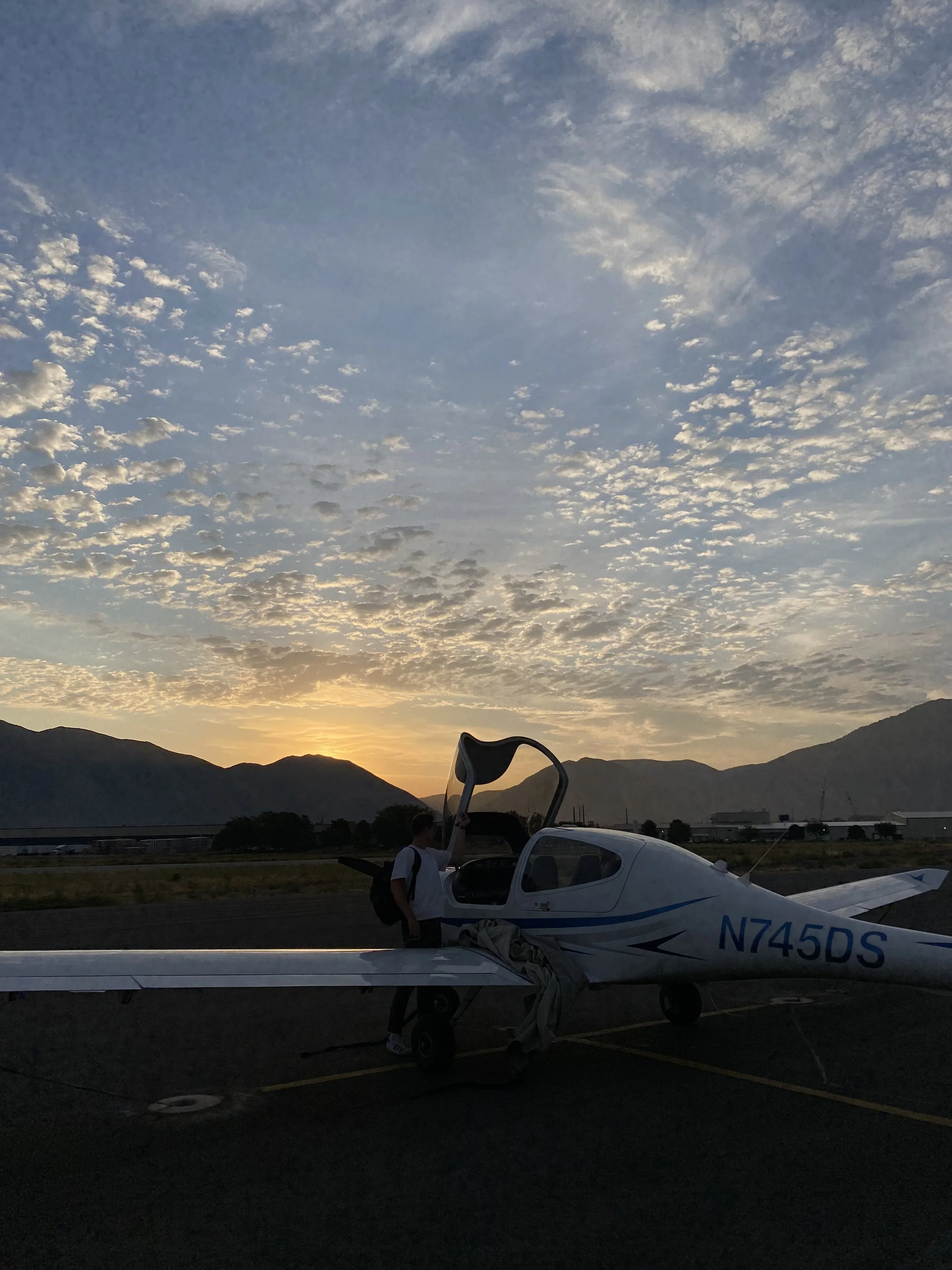 Person standing next to a small white airplane, preparing for flight at sunrise with mountains in the background.