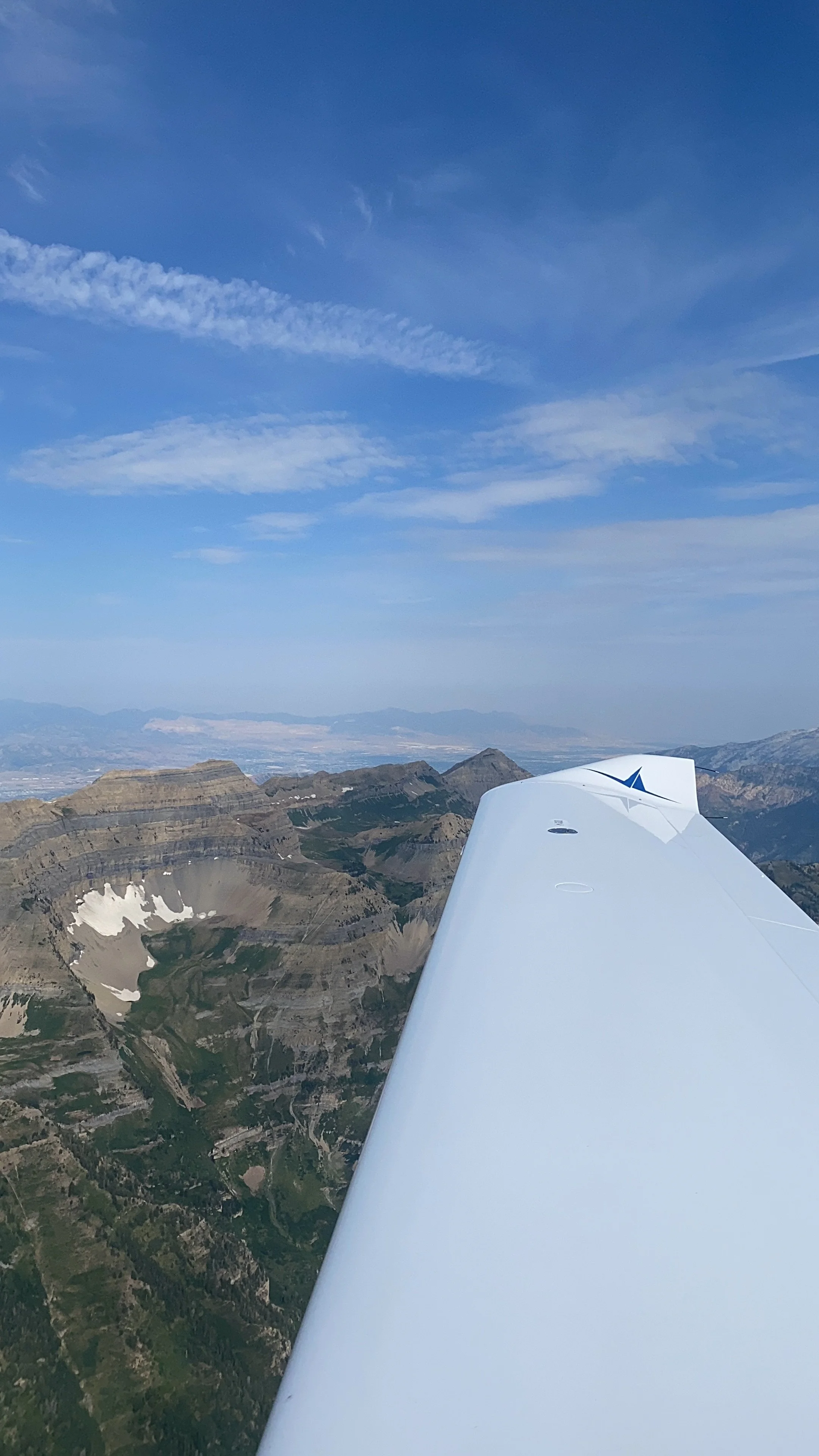 A view from an airplane window showing the wing and a mountainous landscape below, with a blue sky and scattered clouds.