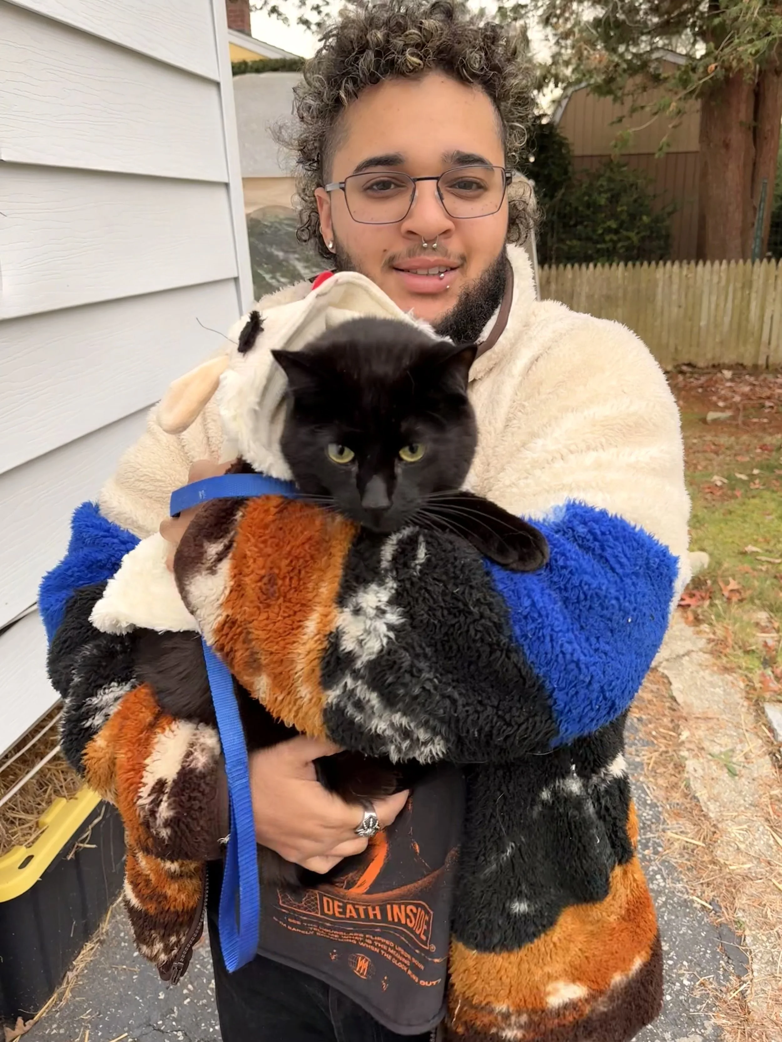 A person with glasses, curly hair, and multiple facial piercings holding a black and white cat dressed in a fluffy costume outdoors. The person is wearing a colorful, plush jacket and a black T-shirt with orange lettering. The background shows a wood