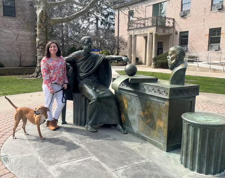 A woman with a dog standing next to bronze statues of three men engaged in a discussion, outdoors in front of a building with trees in the background.