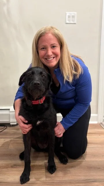 A woman with long blonde hair smiling and crouching next to a black dog with a red collar indoors.