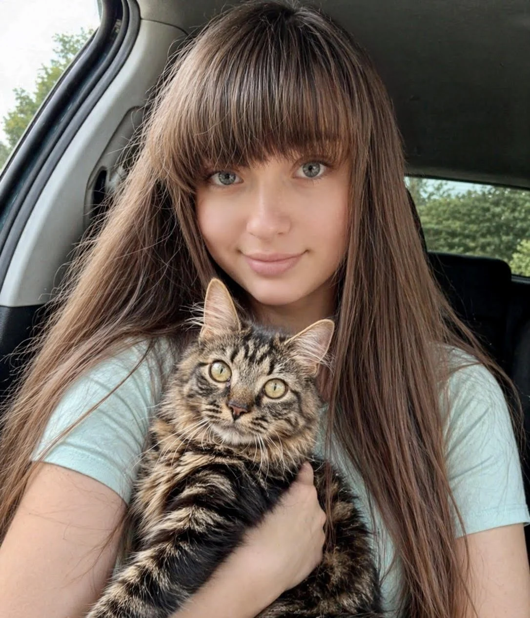 A young woman with long brown hair and blue eyes holding a tabby cat with yellow eyes inside a car.
