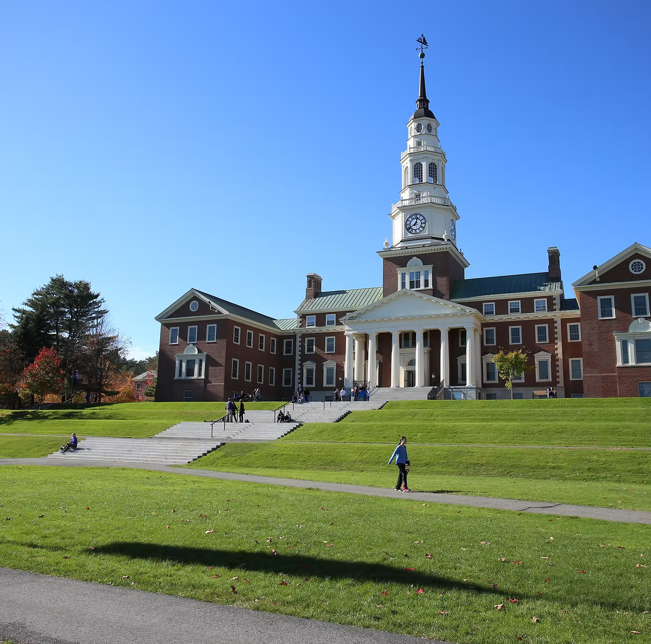 Historic red brick college building with a tall clock tower and a white portico on a sunny day, with students sitting and walking on green grass and stairs.