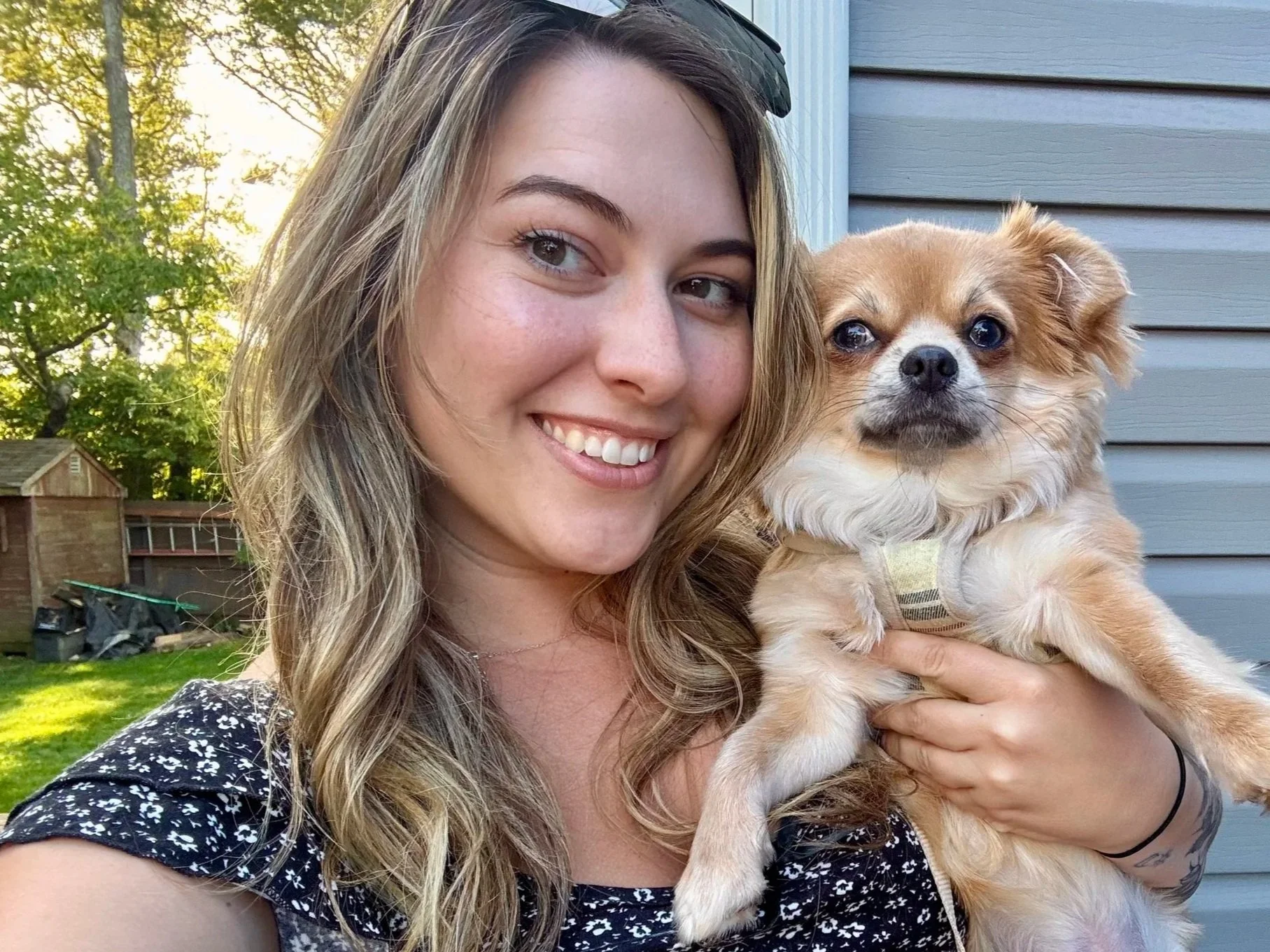 A woman smiling and holding a small tan and white dog in her arms outdoors with trees and a house in the background.