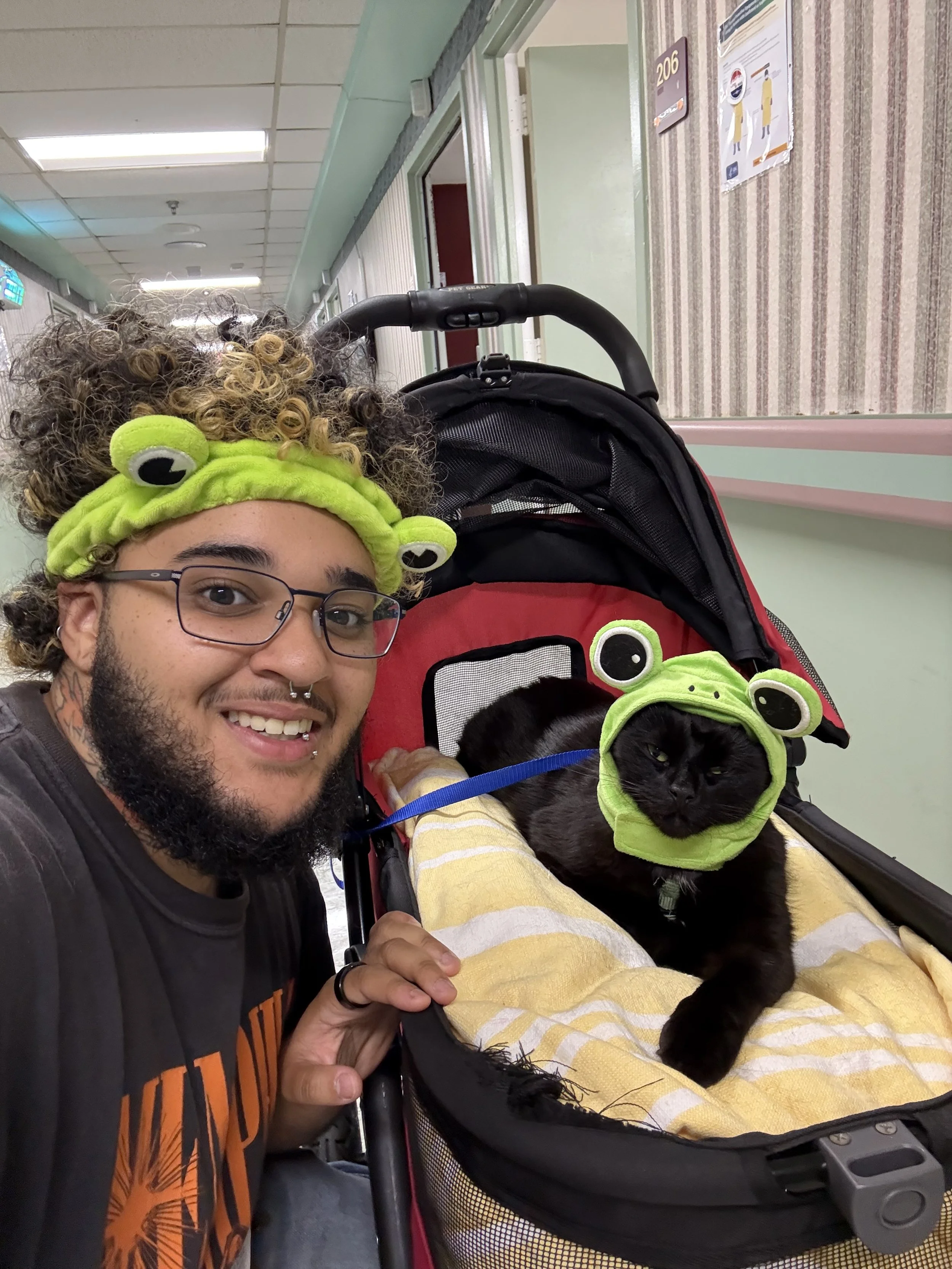 A person with curly hair, wearing a green frog hat, glasses, and a piercings, smiling while taking a selfie with a black dog dressed in matching frog costume, sitting in a stroller in a hospital corridor.