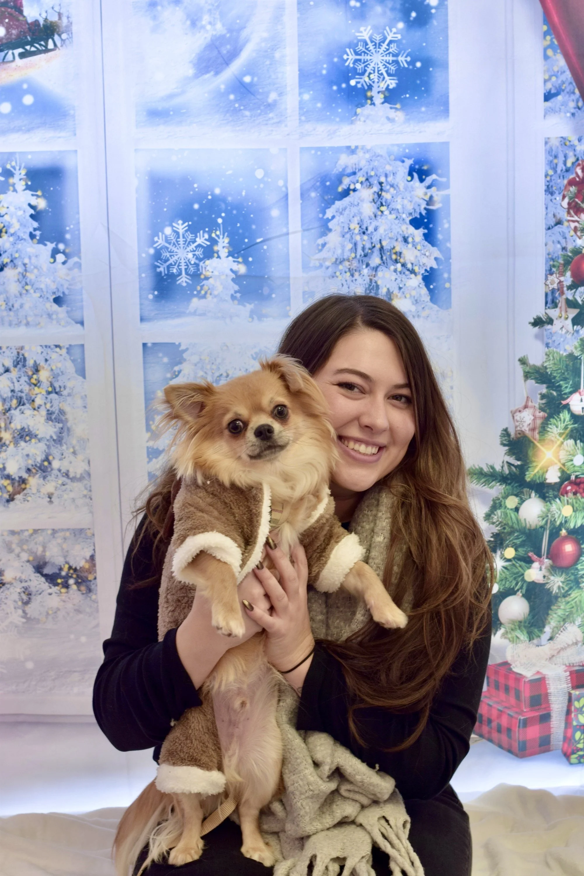 A woman with long brown hair smiling and holding a small tan and white dog in front of a festive winter holiday background featuring snowflakes, snow-covered trees, and presents.