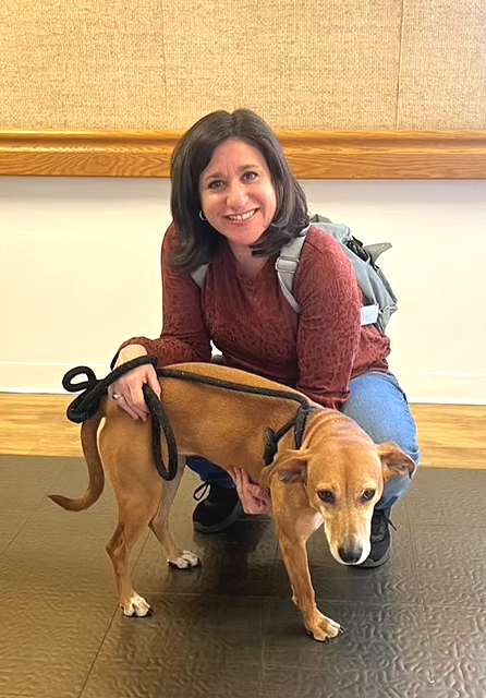 Woman with a backpack crouching next to a brown dog on a leash indoors