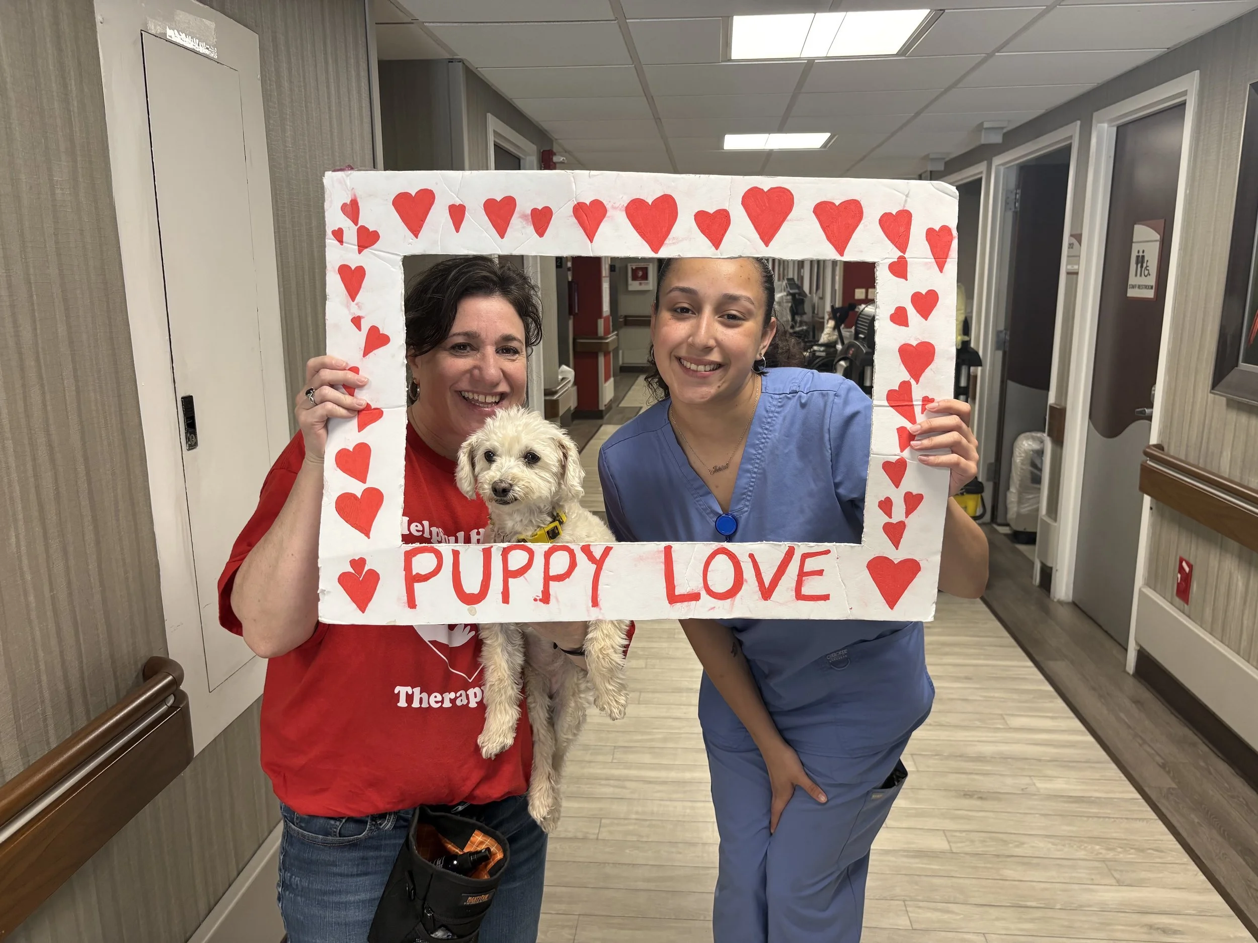 Two women, one in red scrubs and the other in blue scrubs, are holding a cardboard frame decorated with red hearts and the words 'Puppy Love.' They are standing in a hospital hallway with a small puppy.