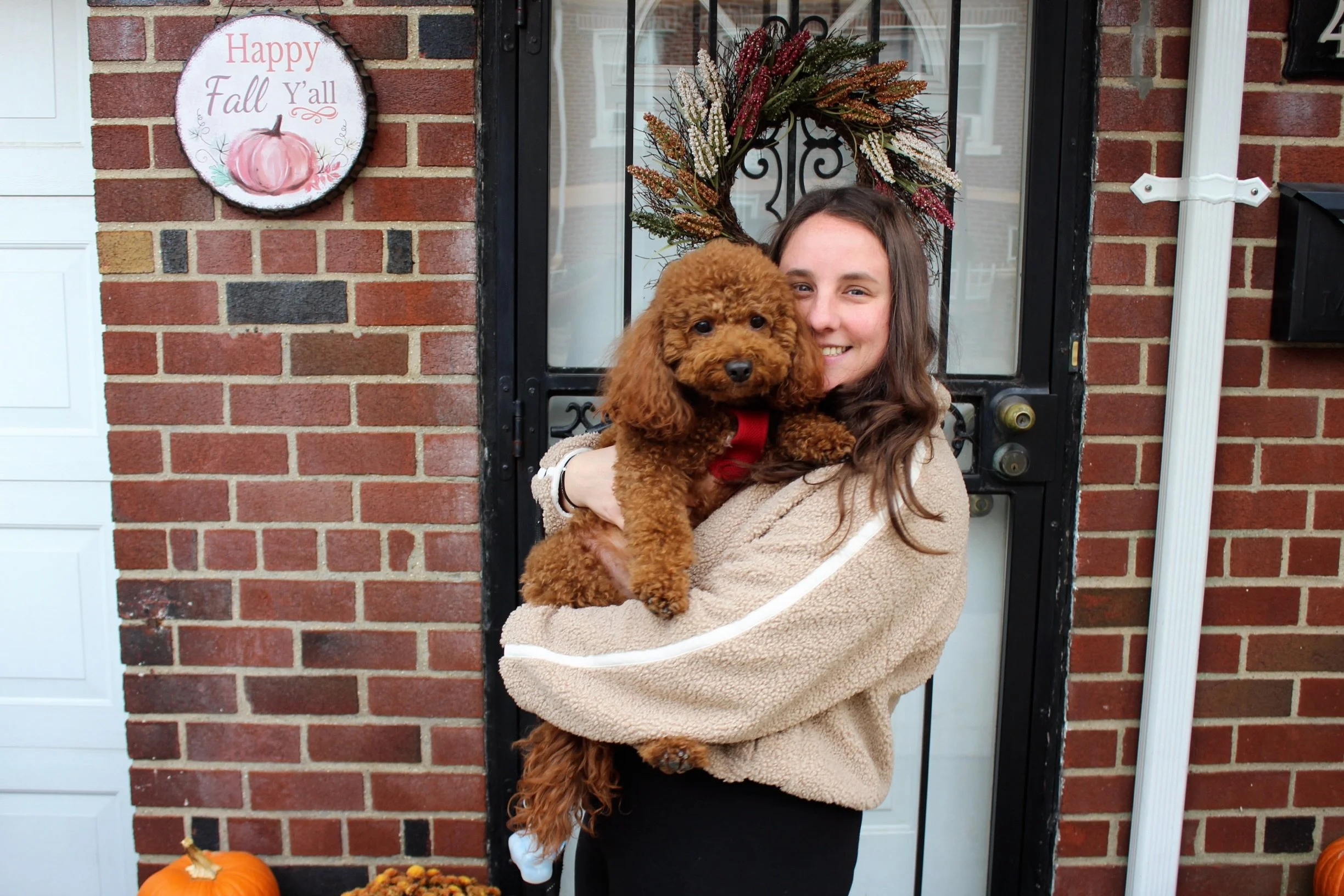 A woman with long brown hair, wearing a beige fleece jacket, holding a small brown poodle puppy with a red harness, standing in front of a brick house entry door decorated with a fall wreath and a fall-themed sign that says 'Happy Fall Y'all' with a pumpkin illustration, surrounded by pumpkins and fall decorations.