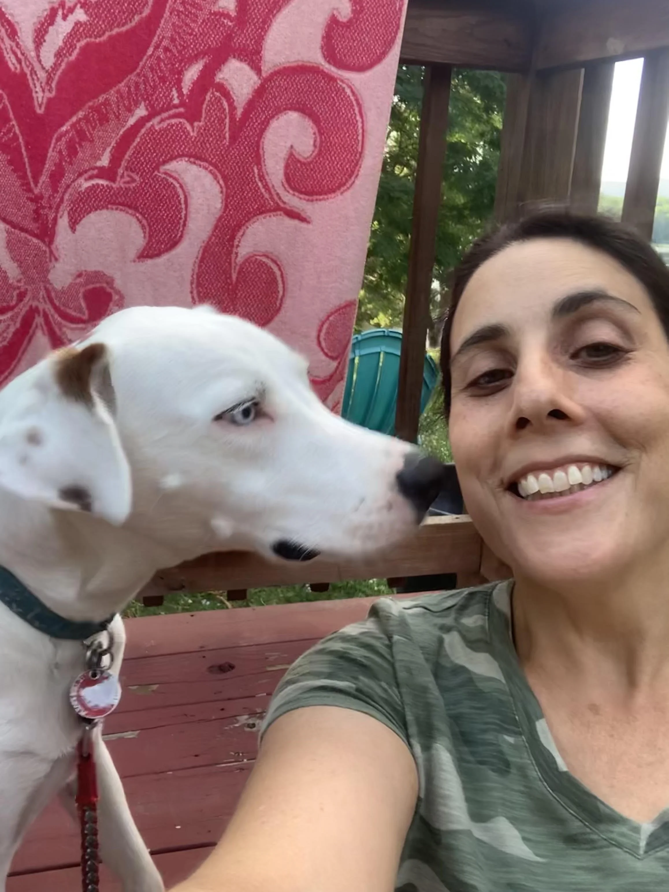 A woman smiling with a dog sitting next to her. The woman has dark hair and is wearing a green camouflage shirt. The dog is white with a brown spots, blue eyes, and is wearing a collar with a red leash. They are outside on a wooden deck with trees and a blue shed in the background.