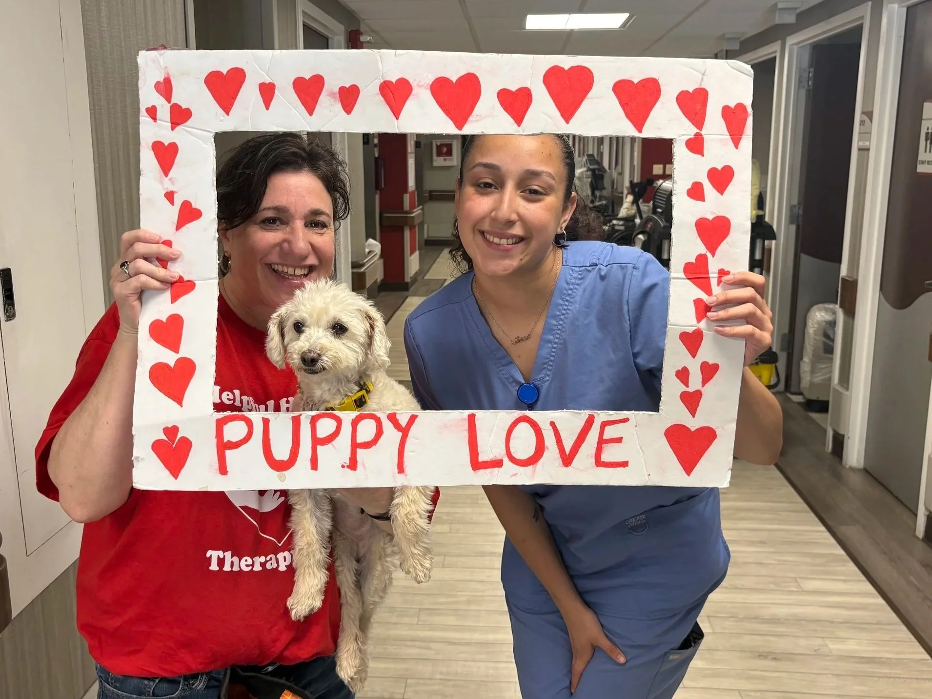 Two women and a dog holding a cardboard frame decorated with red hearts, with the words 'Puppy Love' written on it. The woman on the left is wearing a red shirt, and the woman on the right is in blue scrubs. They are smiling and standing in an indoor facility.