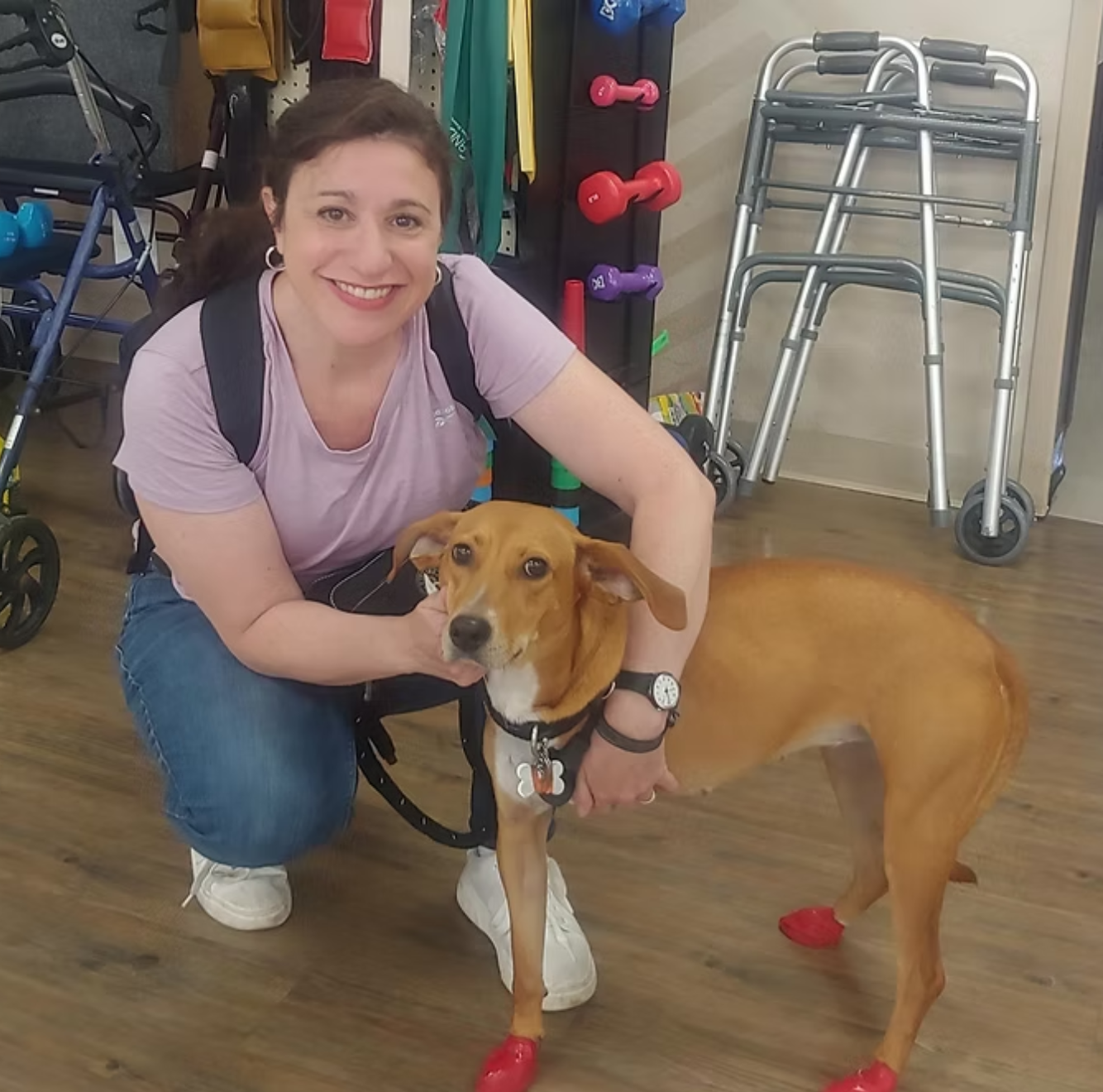 A woman kneeling next to a service dog inside a store or medical facility. The woman is smiling, wearing a light purple T-shirt, jeans, and white shoes. The dog has red boots on its paws and is wearing a harness with a badge. In the background, there are mobility walkers, a shopping cart, and colorful dumbbells.