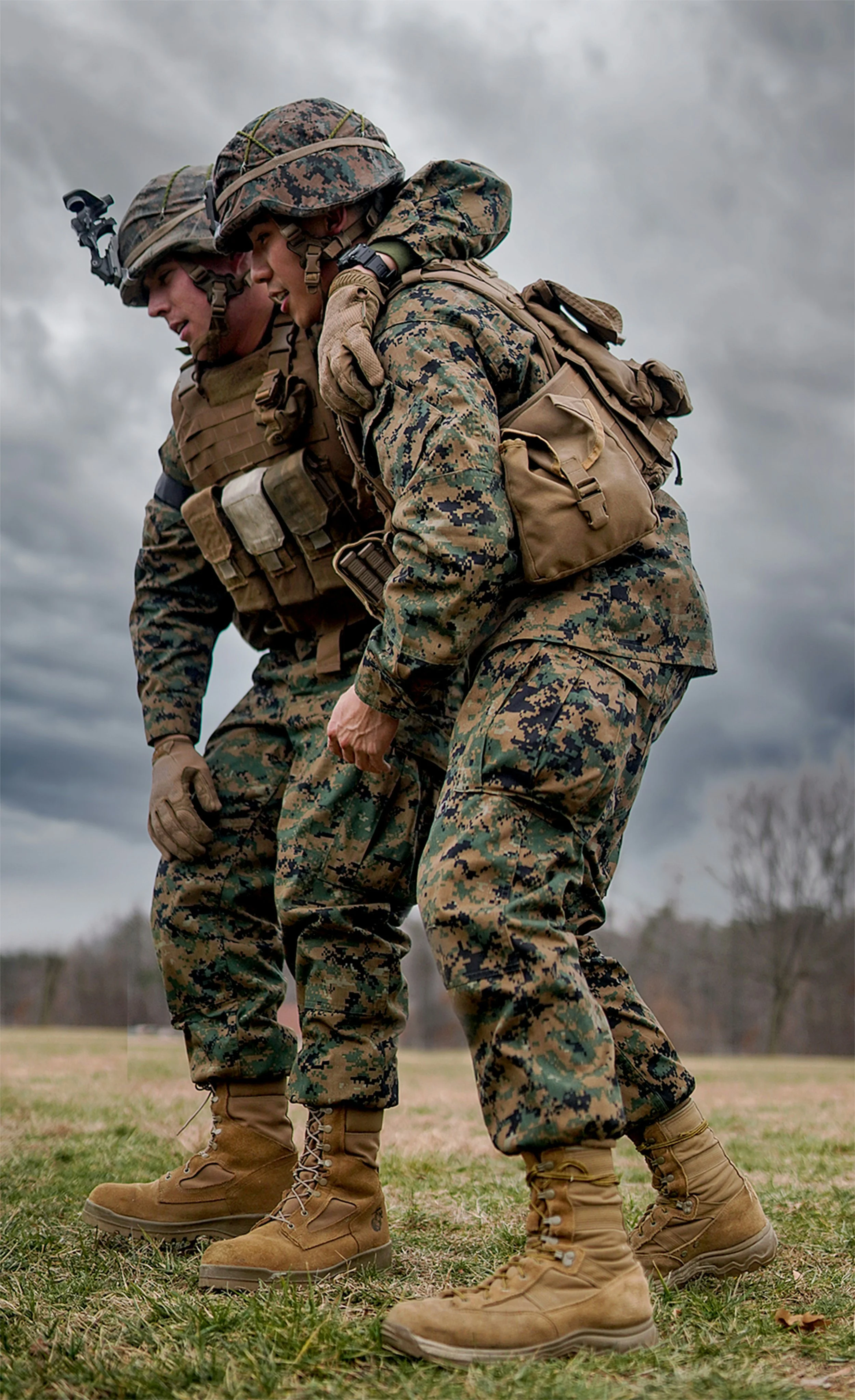 Two soldiers in camouflage uniform assisting each other while standing on a grassy field with cloudy sky.