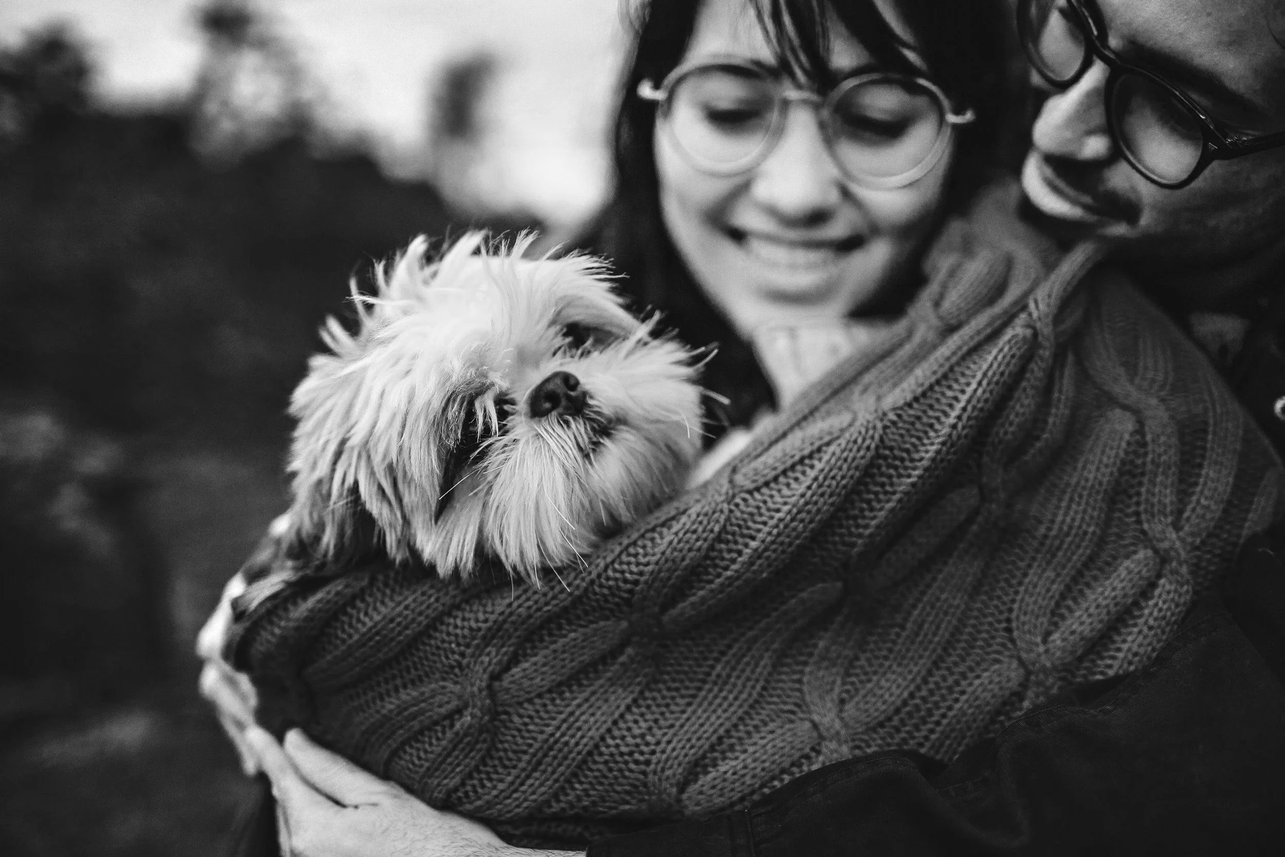 A woman and a man are smiling and holding a small, fluffy dog close to their faces. The woman is wearing glasses and a knitted sweater, the man is also wearing glasses. The image is in black and white.