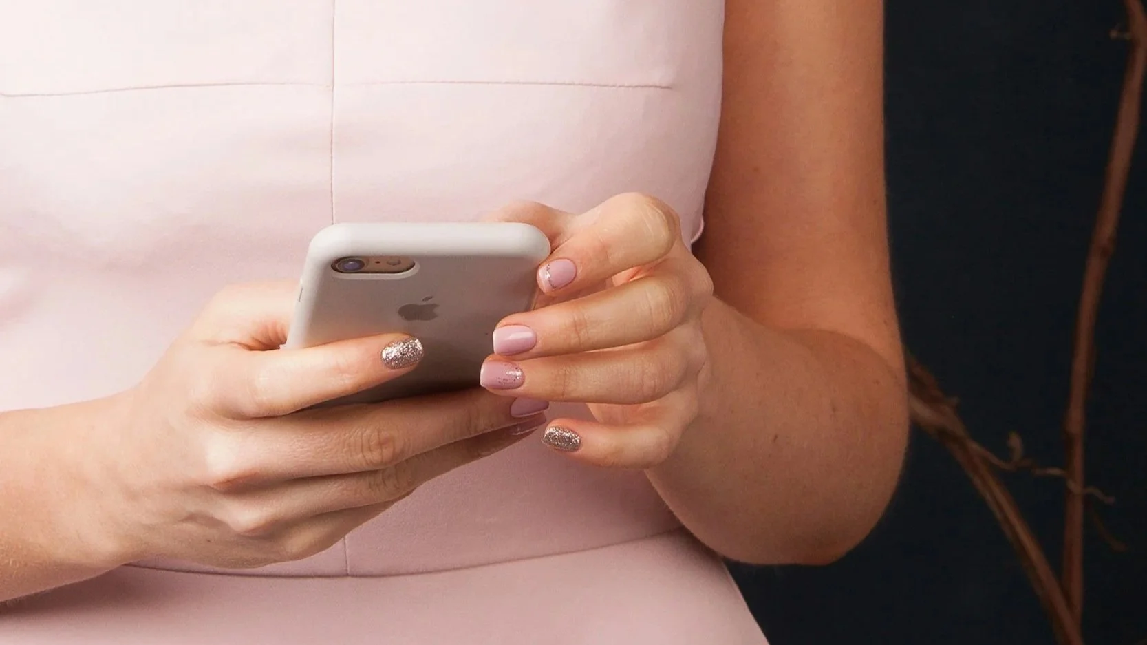 Person holding a white smartphone, wearing a light pink dress with nails painted in pink and glitter, with a dark background.