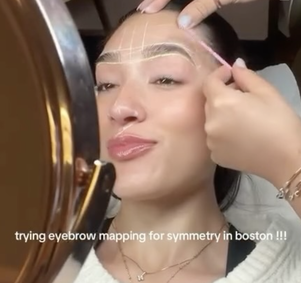 A woman is getting her eyebrows mapped for symmetry with dotted lines drawn above her eyebrows. A beautician is using a pink tool to mark her eyebrows while the woman relaxes at a beauty salon mirror. The caption reads, 'trying eyebrow mapping for symmetry in boston !!!'