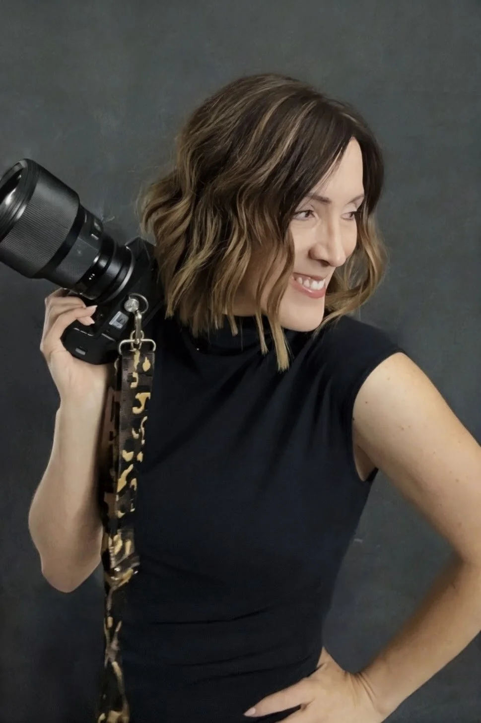 A woman with wavy brown hair smiling, holding a camera on her shoulder, wearing a black sleeveless top, against a dark background.