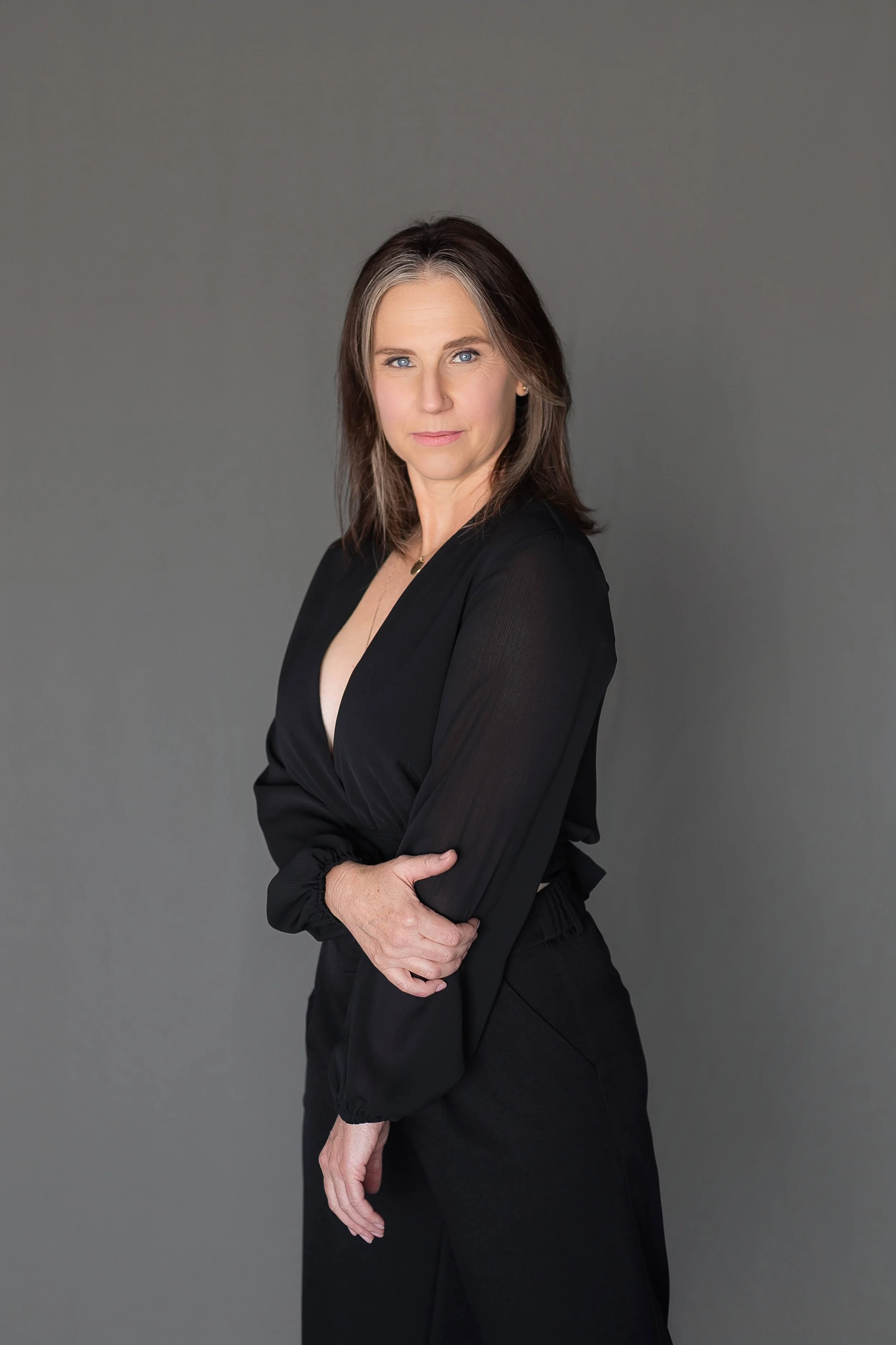 A woman with shoulder-length brown hair and blue eyes wearing a black long-sleeve top, standing against a plain gray background.