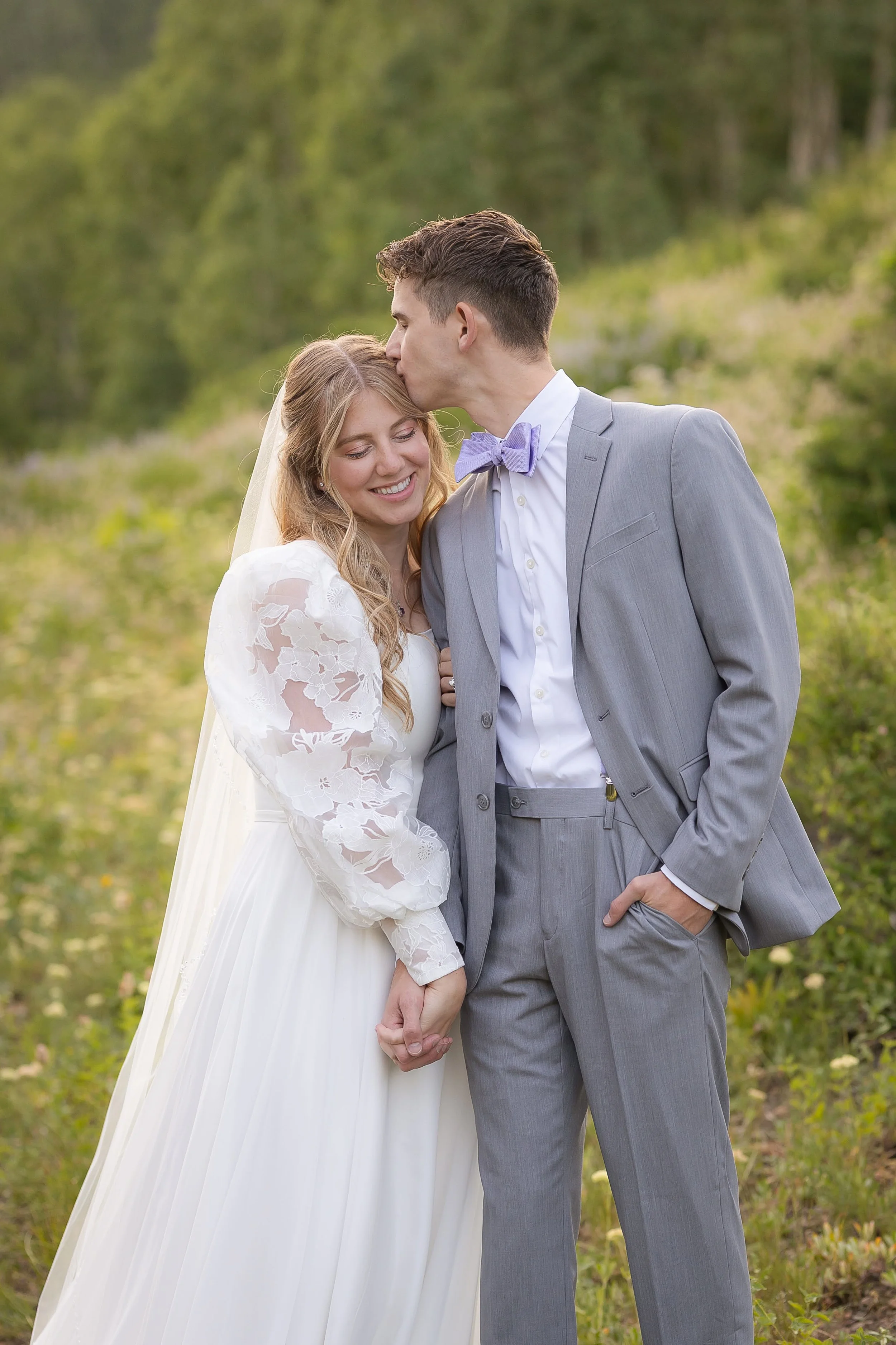 A bride and groom holding hands, with the groom kissing the bride's forehead, outdoors with greenery in the background.