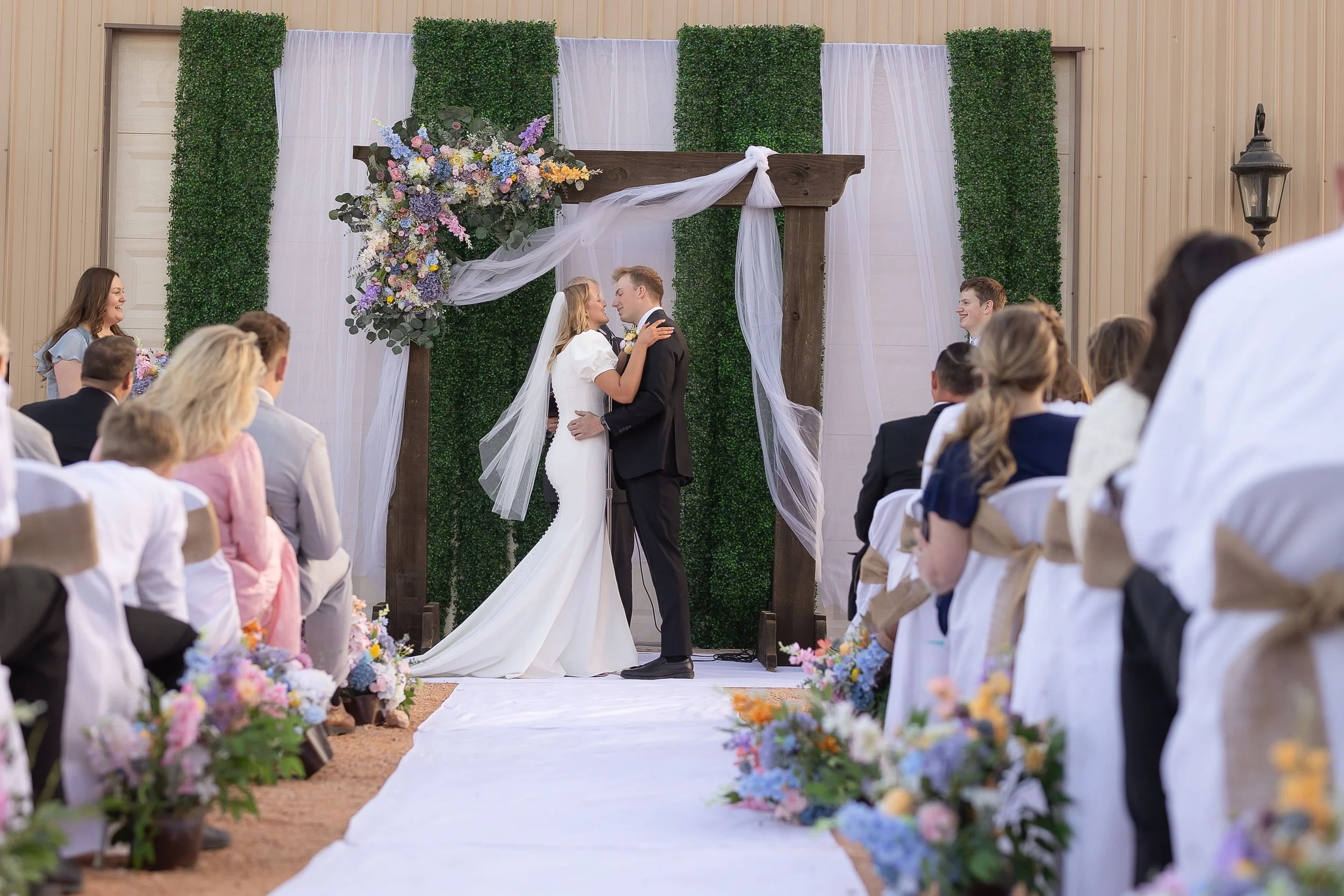 A bride and groom standing close together under a decorated wedding arch, surrounded by seated guests, during a wedding ceremony.