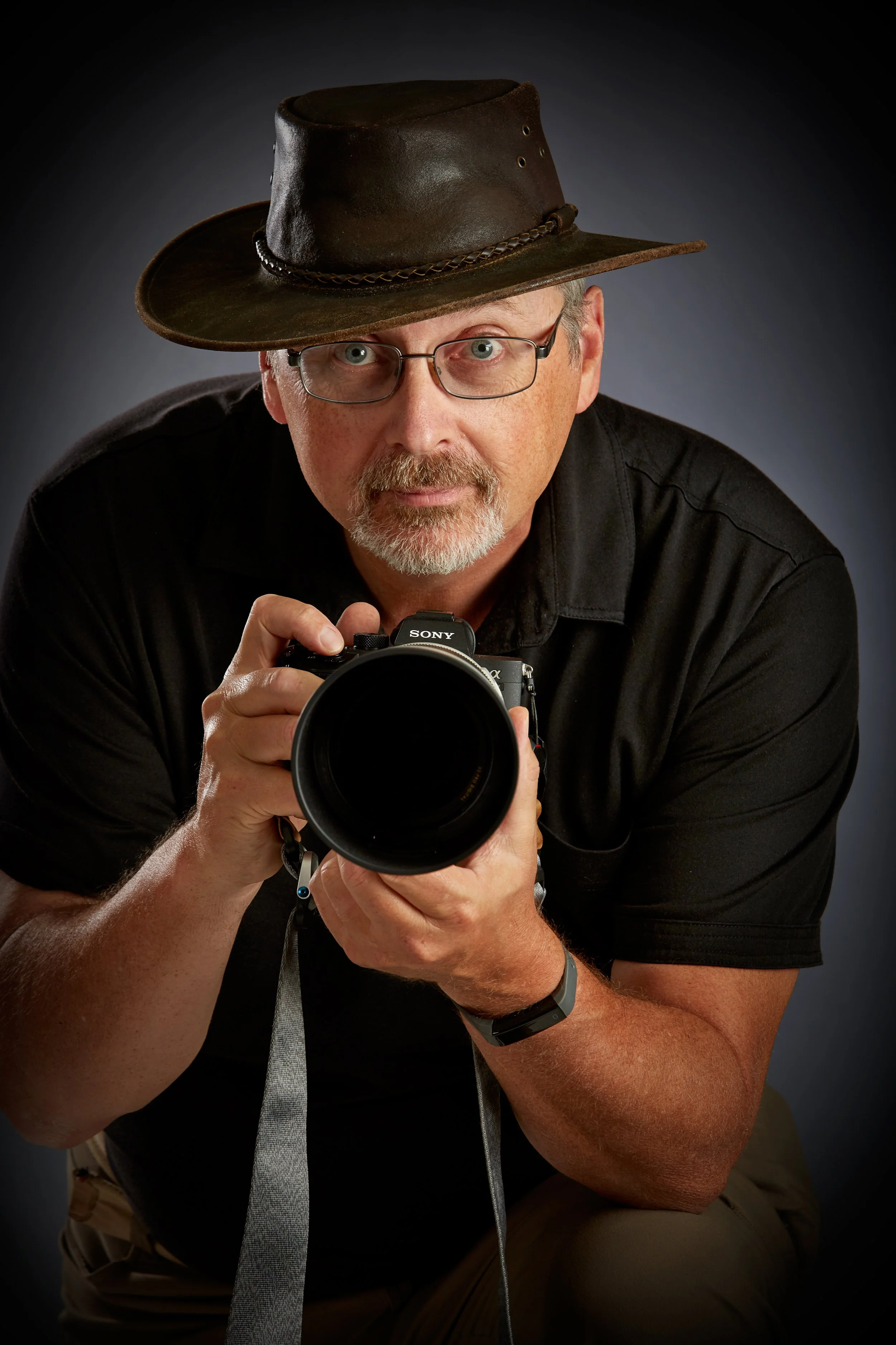 A man wearing glasses, a cowboy hat, and a black shirt holding a Sony camera, looking directly at the camera with a dark background.